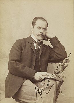 Stare zdjęcie przedstawia mężczyznę w średnim wieku, ma krótkie włosy, wysokie czoło i wąs. Lewą ręką podtrzymuje głowę opierając się łokciem o oparcie krzesła. W prawej ręce trzyma książkę. Na zdjęciu widnieje podpis.