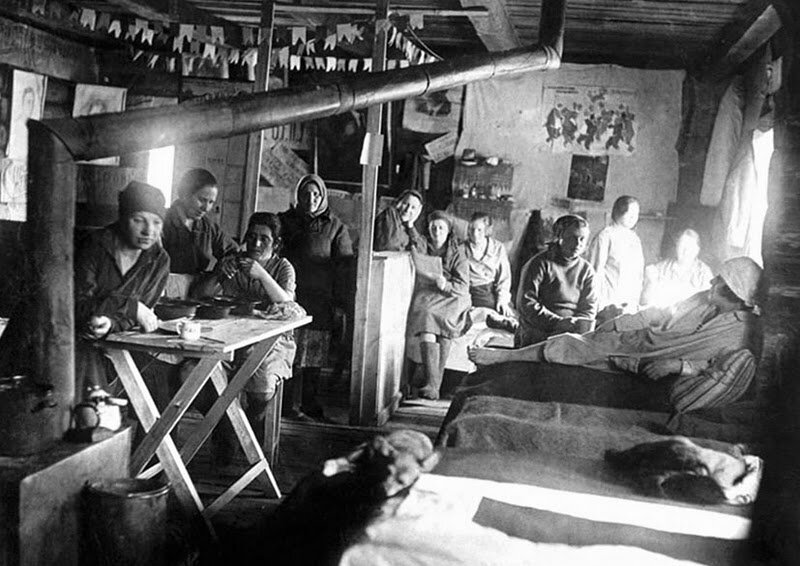 Czarno‑białe zdjęcie przedstawia pomieszczenie z ustawionymi łózkami po prawej oraz drewnianym stołem i piecem z biegnącą do sufitu rurą po lewej. W pomieszczeniu znajduje się grupa jedenastu kobiet w różnym wieku. Ubrane są w proste suknie, mają na głowach chusty. Kilka siedzi przy stole, pozostałe siedzą na łóżkach, a jedna leży. Na ścianie w tle wiszą rysunki i plakaty.