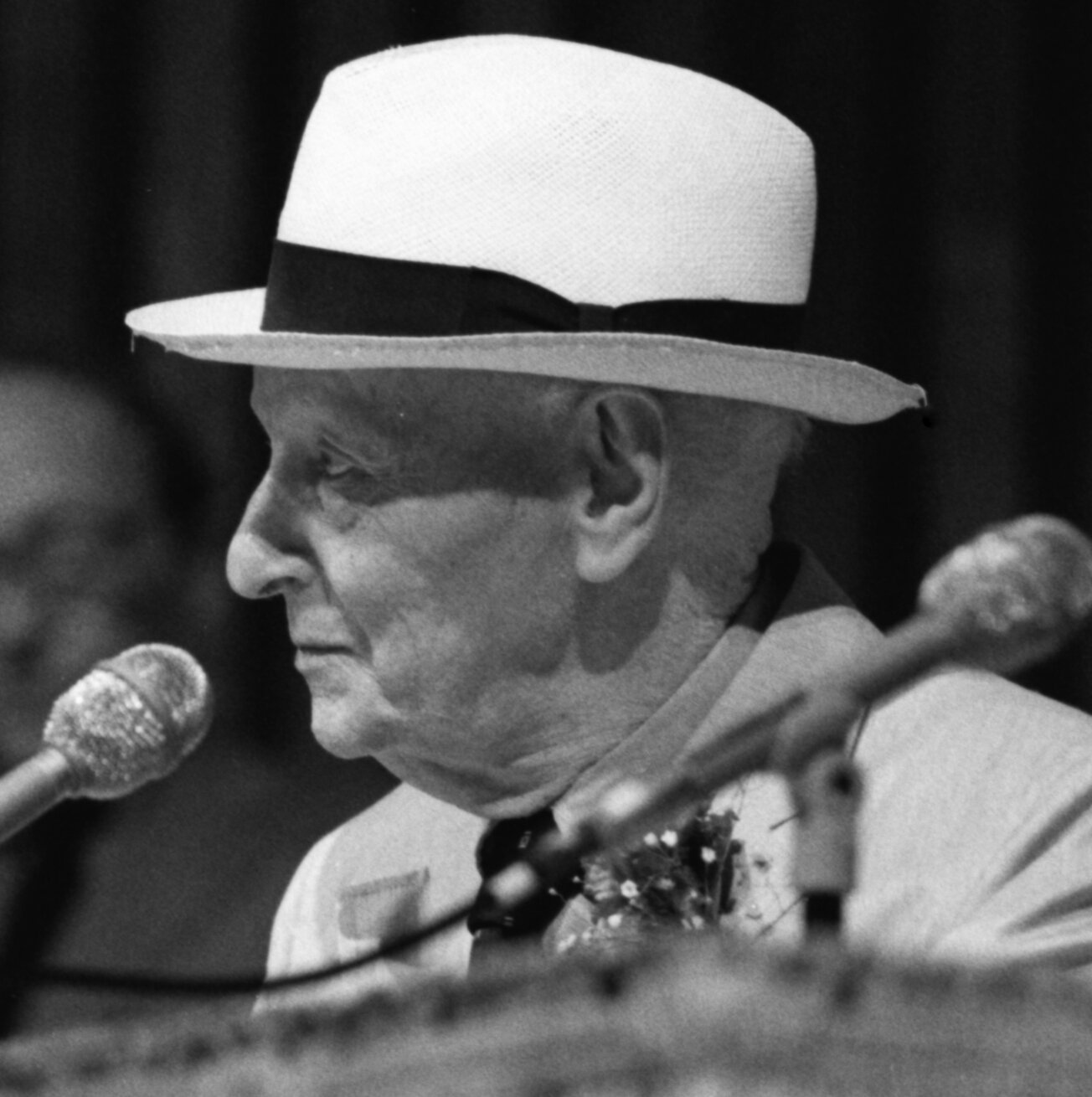 Zdjęcie przedstawia portret starszego mężczyzny w jasnym garniturze i białym kapeluszu, który trzyma gitarę (widoczny gryf instrumentu). Przed nim znajduje się mikrofon. Tło jest ciemne.