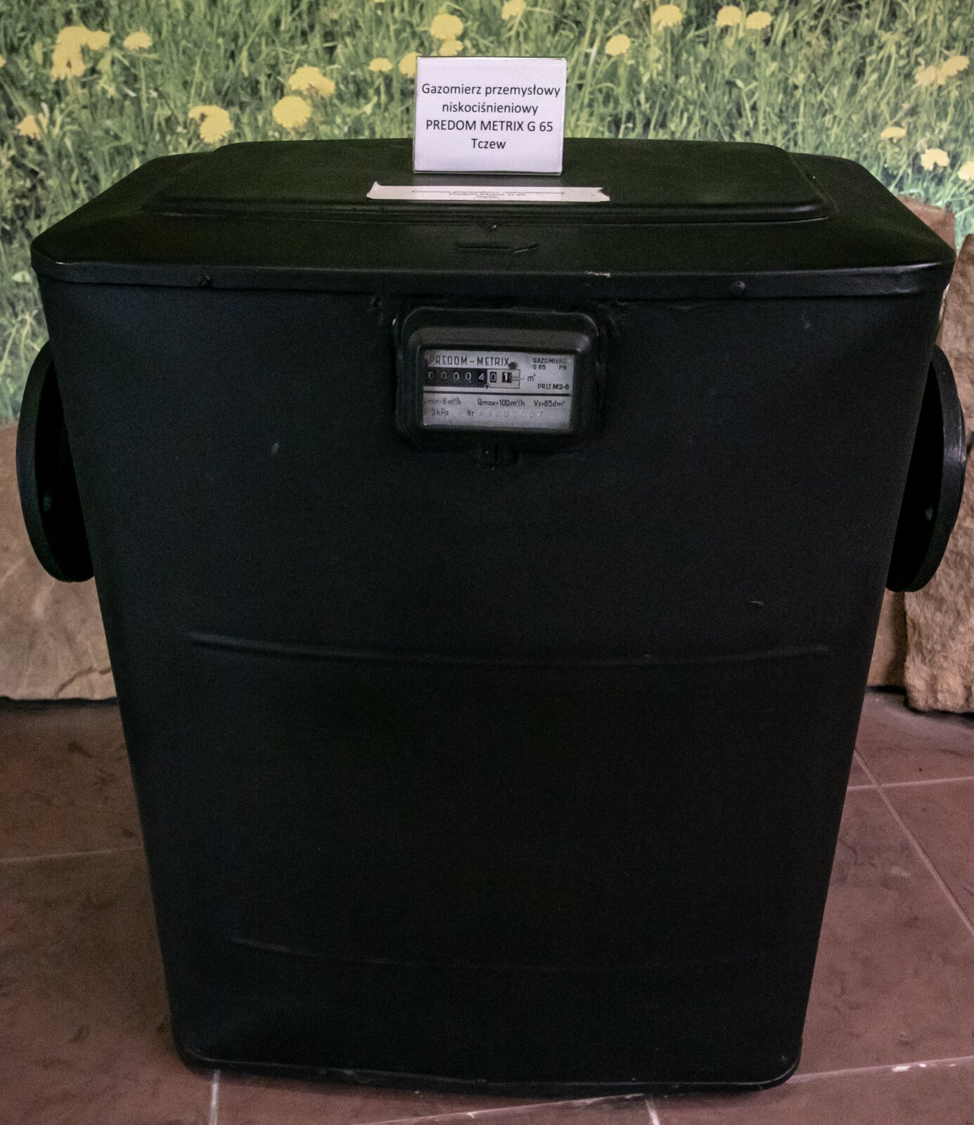 Zdjęcie przedstawia gazomierz przemysłowy niskociśnieniowy PREDOM METRIX G 65 Tczew. Gazomierz jest koloru czarnego. Wyglądam przypomina duże pudełko. Na jego szczycie stoi tabliczka z nazwą. Na przedniej obudowie znajduje się okienko z licznikiem. Na prawej i lewej ściance obudowy znajdują się okrągłe uchwyty.