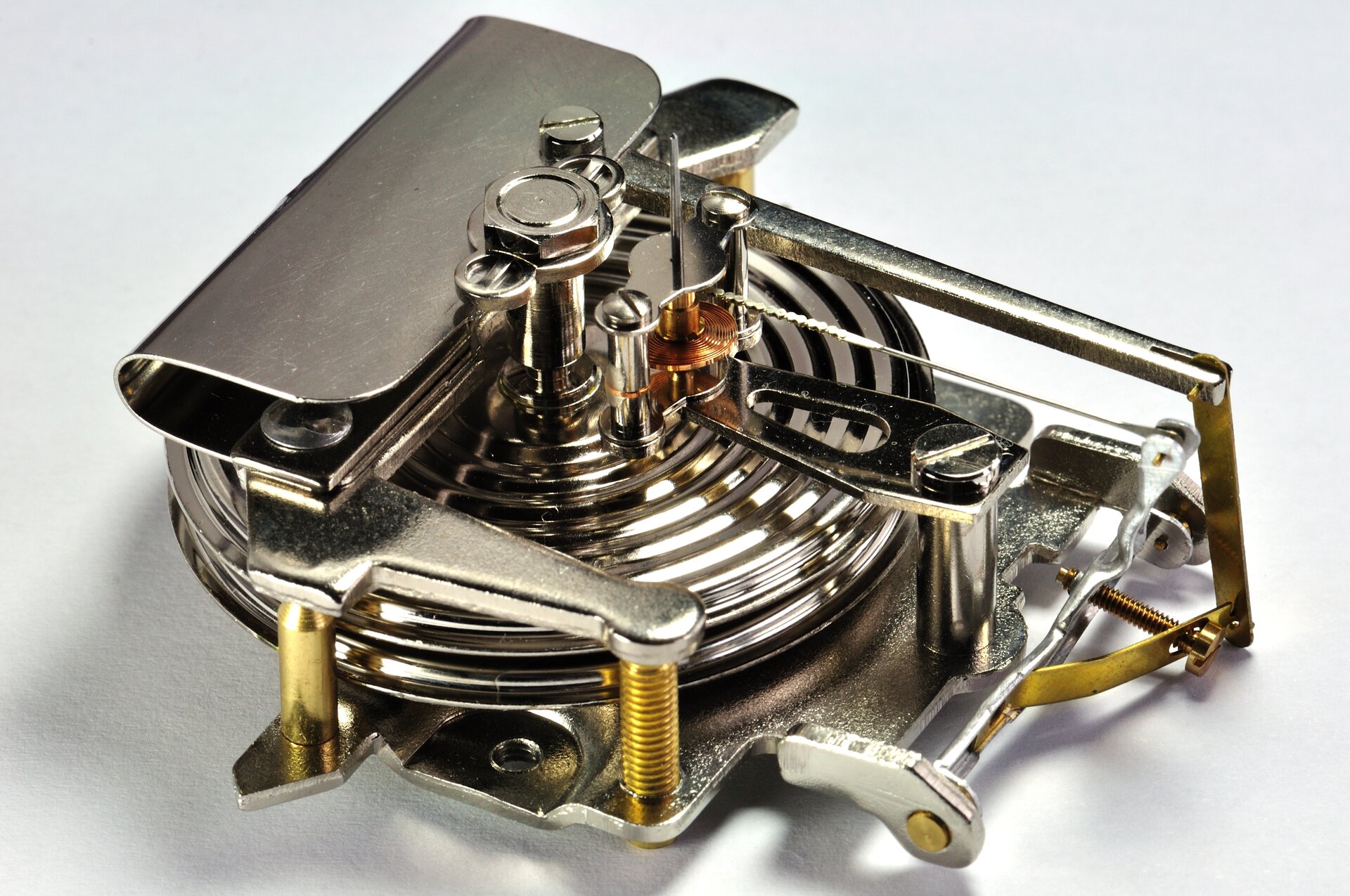 Zdjęcie przedstawia wnętrze aneroidu. Tło białe. Urządzenie wykonane z metalu. Wewnątrz skomplikowanego mechanizmu wyraźnie widoczna puszka Vidiego o kształcie spłaszczonego walca o pofałdowanych podstawach.