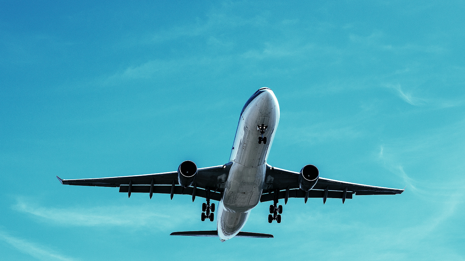 Zdjęcie przedstawia lecący samolot pasażerski na tle bezchmurnego nieba, z perspektywy osoby obserwującej z ziemi. Widoczne jest opuszczone podwozie samolotu.