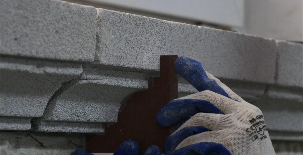 Zdjęcie przedstawia przeciwszablon (kontrszablon), który jest przykładany przez pracownika do gzymsu budynku. Kontrszablon odpowiada kształtowi gzymsu. Na fotografii widoczna jest dłoń pracownika w rękawicy ochronnej.