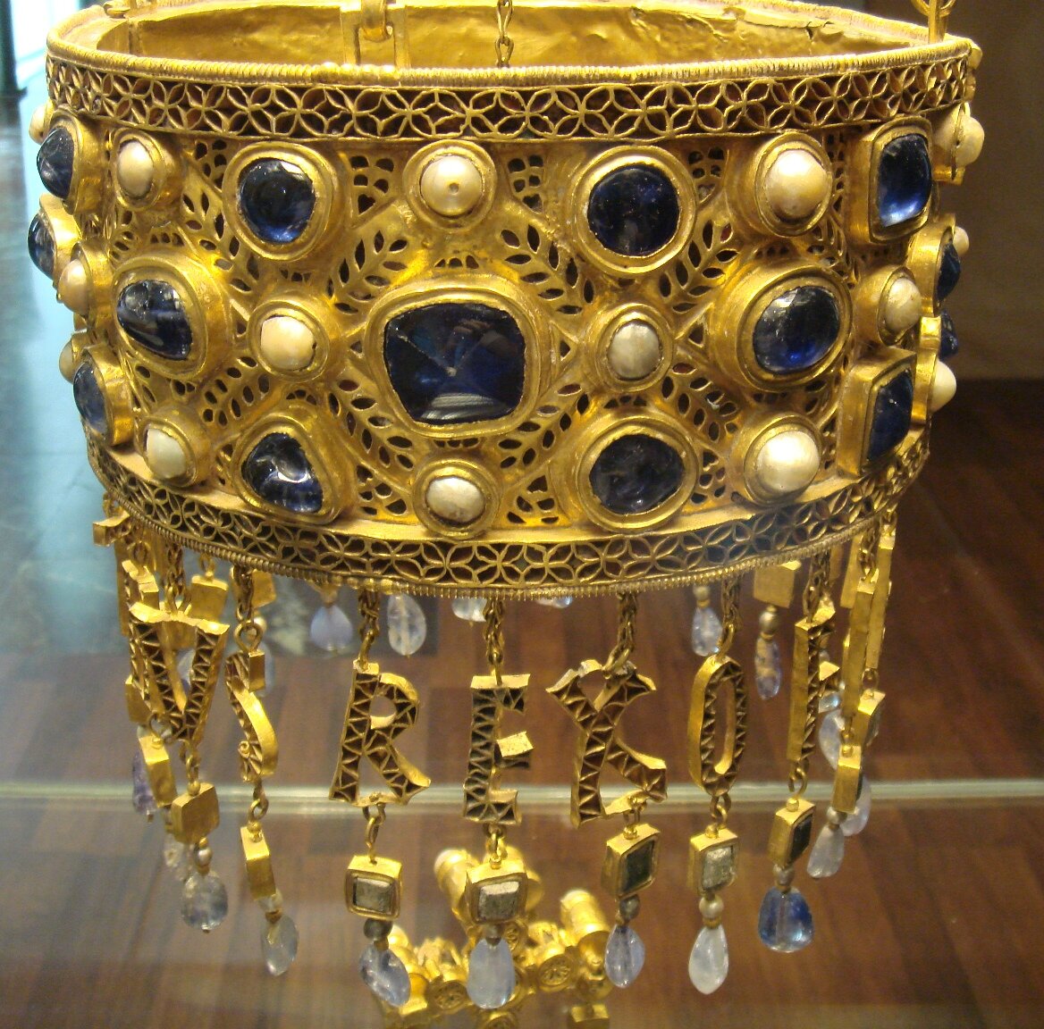 Ilustracja przedstawia złotą koronę ze szlachetnymi kamieniami ułożonymi symetrycznie, w kolorach błękitnym i białym. Pod koroną podwieszane na złotych łańcuszkach kryształy i wykonane ze szlachetnego kamienia litery. Widok współczesny. 