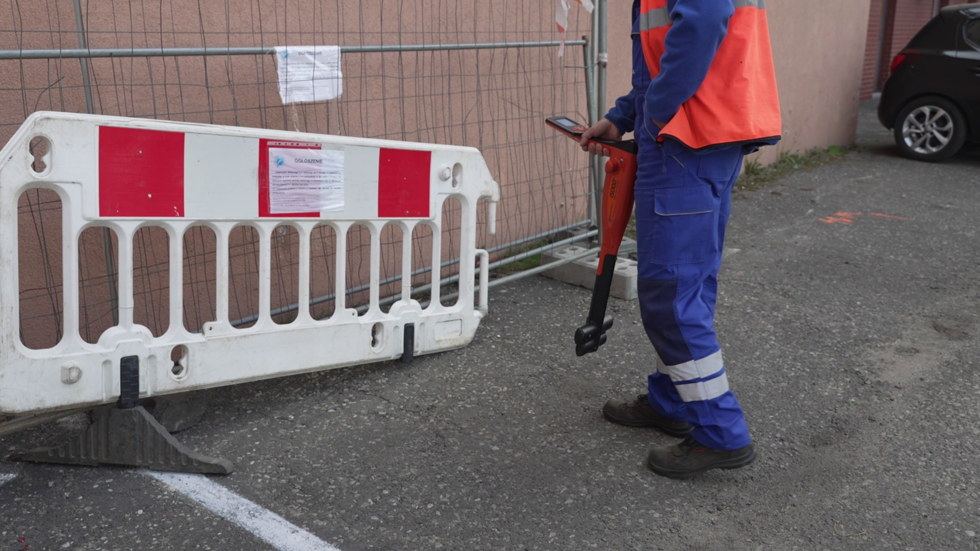 Na ekranie mężczyzna ubrany w strój roboczy trzymający w rękach urządzenie do pomiaru. W tle auto i budynek, a po lewej stronie bariera oddzielająca.