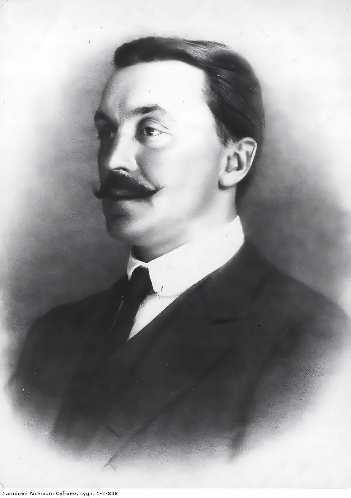 Zdjęcie przedstawia portret mężczyzny w średnim wieku. Mężczyzna ma ciemne włosy zaczesane do tyłu. Ma ciemne oczy i długi, prosty nos. Ma ciemny wąs, który jest podkręcony na końcach. Ubrany jest w białą koszulę, czarny krawat i marynarkę. Ciało mężczyzny jest skierowane w lewą stronę zdjęcia - w tym samym kierunku również patrzy. Zdjęcie jest czarno‑białe.