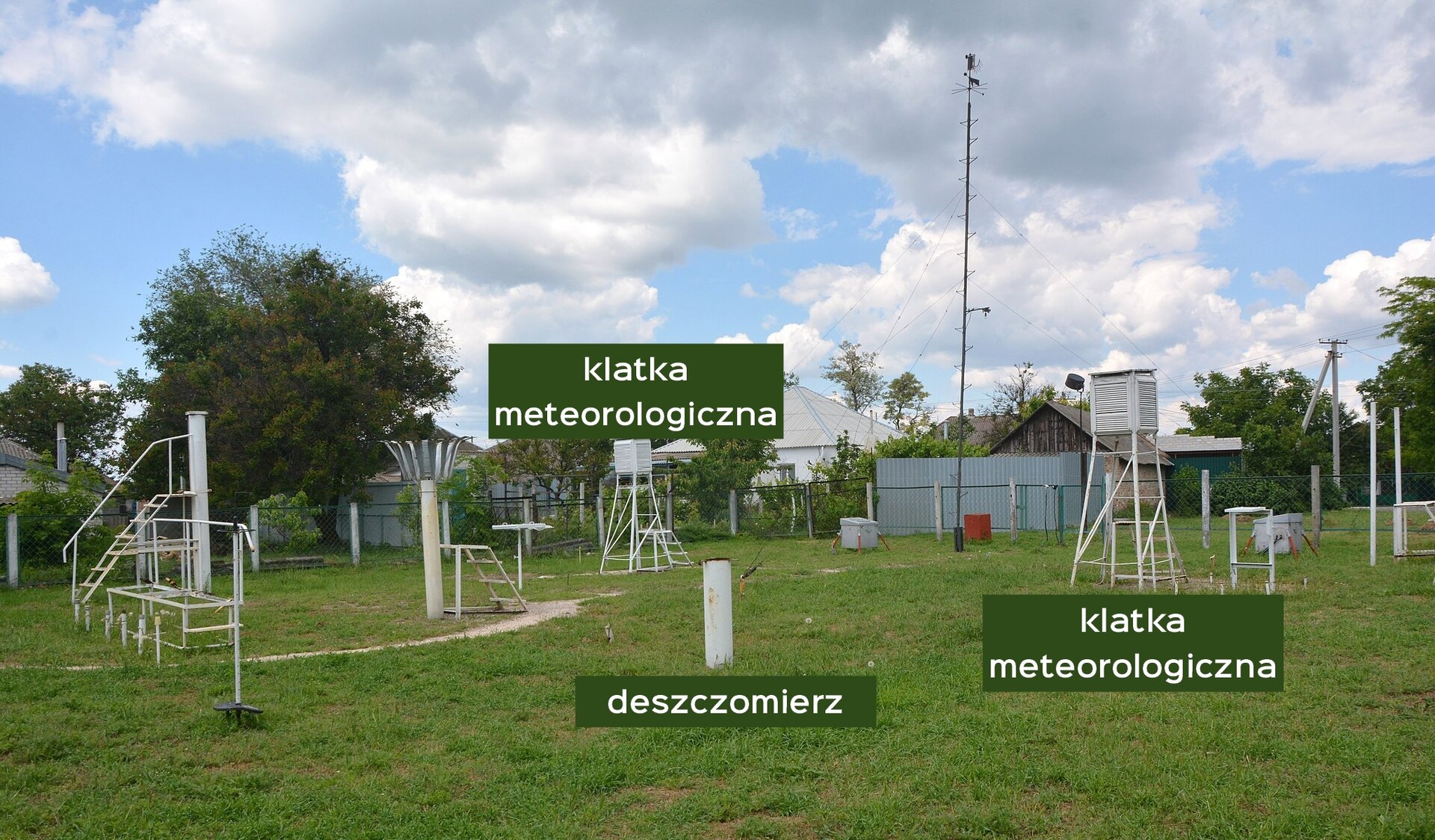 Zdjęcie przedstawia przykładowy ogródek meteorologiczny. Widoczne są dwie klatki meteorologiczne i jeden deszczomierz. Deszczomierz z boku wygląda jak walec wystający pionowo z ziemi. Klatki meteorologiczne stoją na pewnym podwyższeniu. W ogródku widoczne są też inne przyrządy o różnorodnych kształtach.