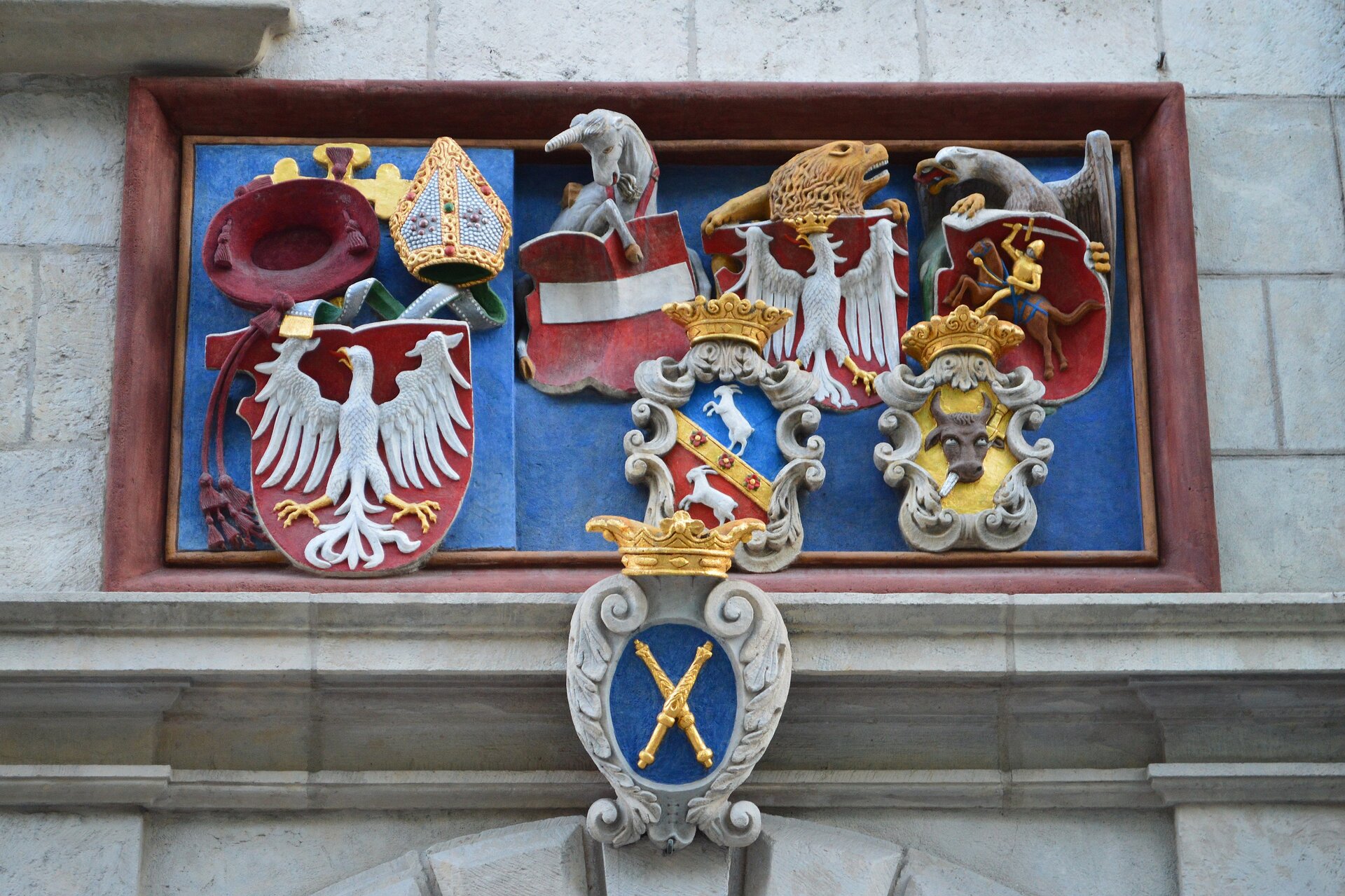 Zdjęcie przedstawia pas dekoracyjny nad wejściem do budynku, na którym to fryzie znajdują się liczne herby. Od lewej herb na czerwonym tle, biały orzeł z głowa zwróconą w prawą stronę, nad tym herbem wydłużona czapka, składająca się z dwóch wysokich, połączonych ze sobą trójkątów, tak zwana mitra. W centralnym miejscu jednorożec przytrzymujący kopytem czerwony herb z białym, poziomym pasem. Poniżej herb zwieńczony złotą koroną, przedstawiający w dolnym - czerwonym i górnym - niebieskim polu dwa koziołki stojące na tylnych nogach. Nad nim czerwony herb z białym orłem w złotej koronie. Z lewej trzymany w szponach orła herb, na którym widnieje jeździec na koniu, z uniesionym w górę mieczem. Poniżej fryzu zdobienie architektoniczne, które przedstawia dwa skrzyżowane berła na niebieskim tle, zwieńczone złotą koroną. 
