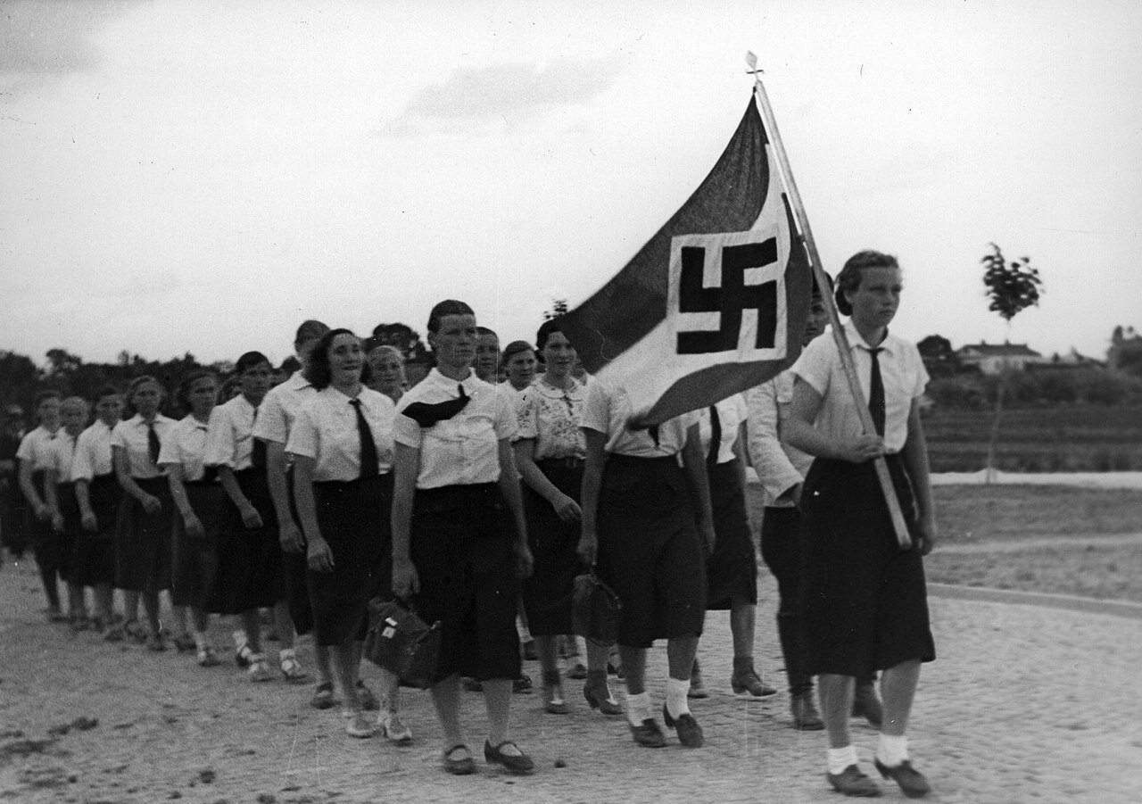 Zdjęcie przedstawia dziewczęta idące w pochodzie, na czele którego stoi dziewczyna z flagą ze swastyką. Dziewczęta ubrane są w czarne spódnice do połowy łydki, białe bluzki i czarne krawaty.