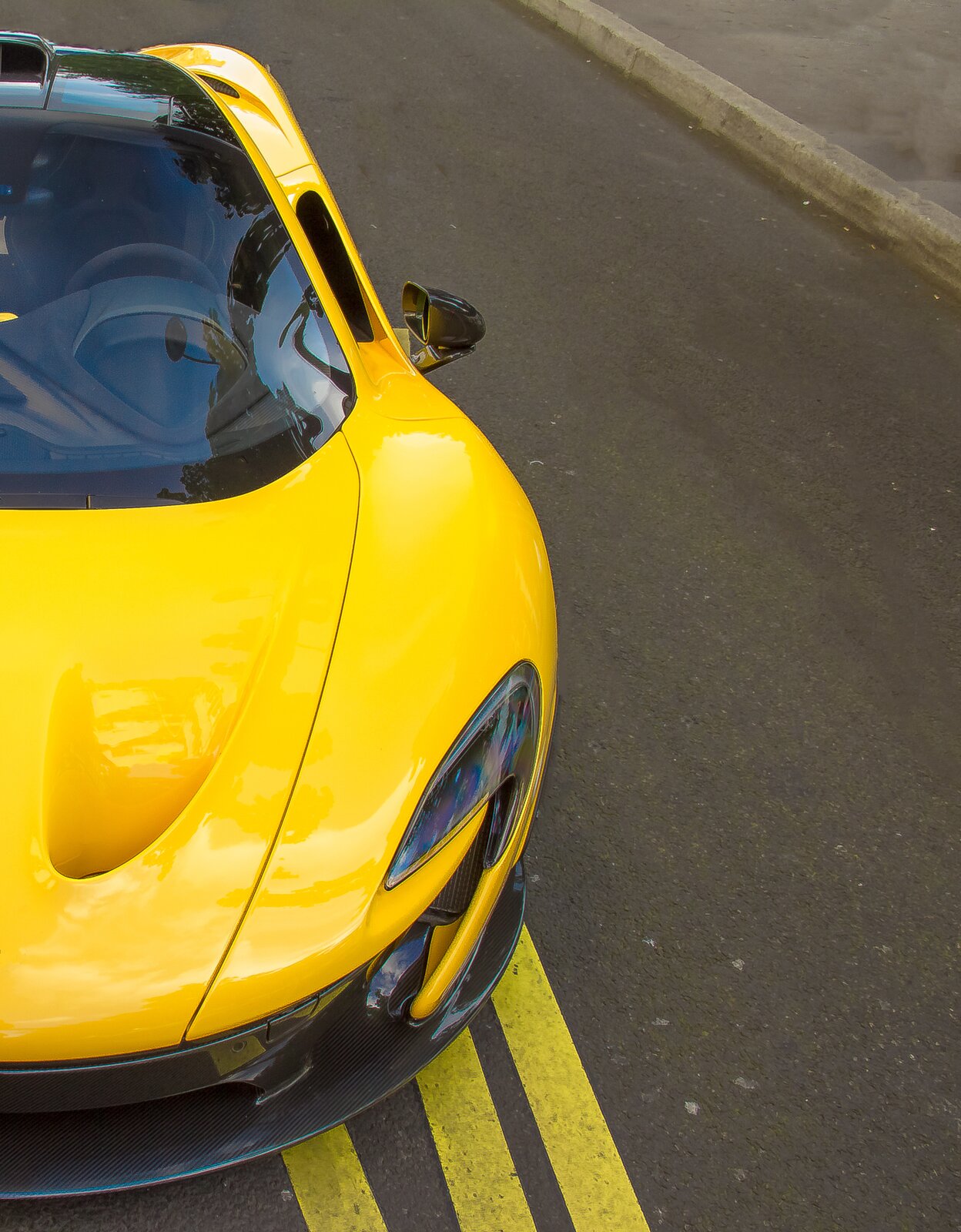 Zdjęcie przedstawia fragment sportowego samochodu. Samochód koloru żółtego, błyszcząca karoseria. Stoi na szarej, asfaltowej drodze.
