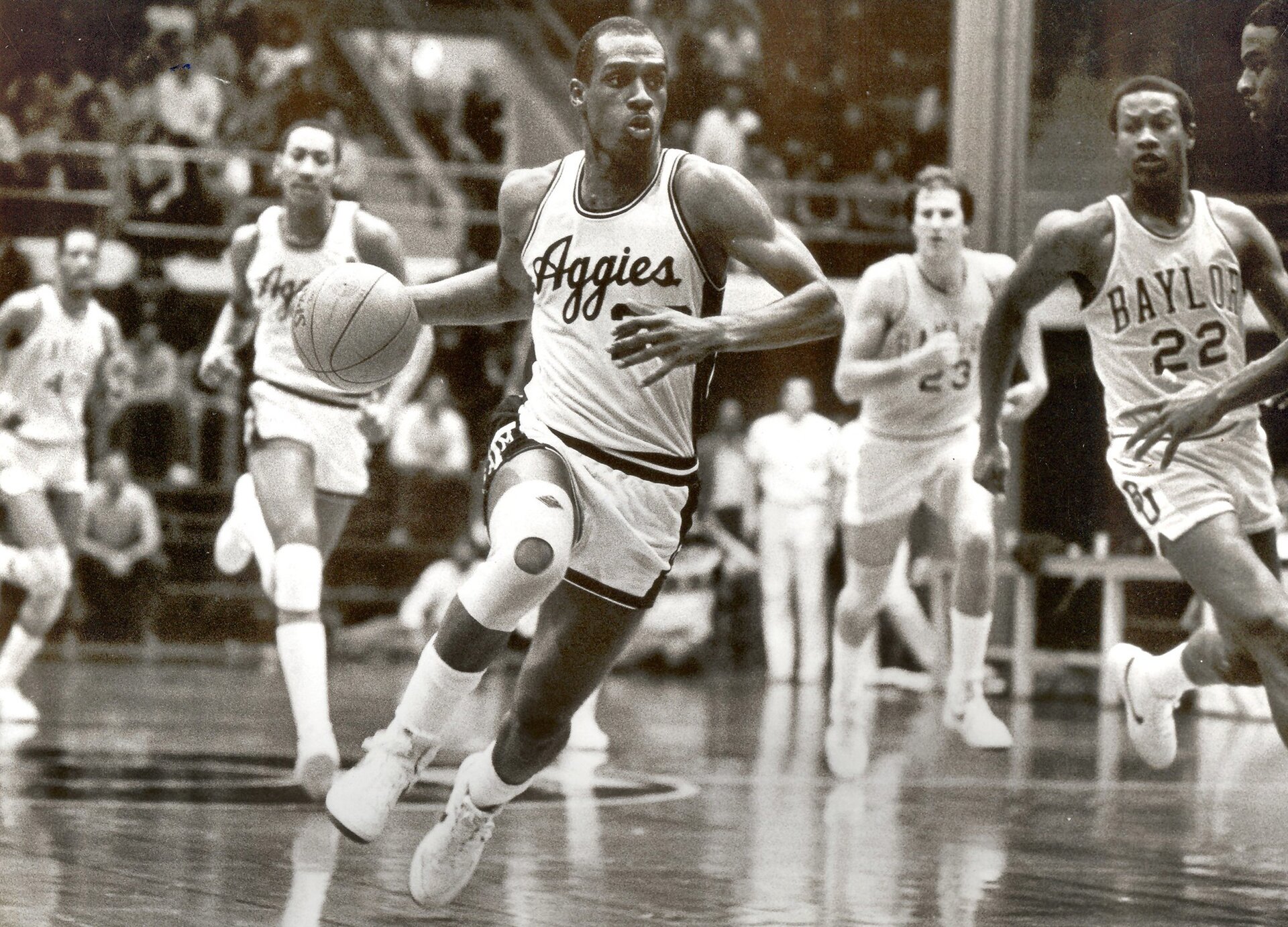 Czarno białe zdjęcie przedstawia mężczyzn grających w koszykówkę na boisku na hali. 