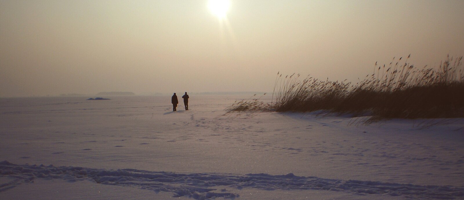 Fotografia przedstawia płaski teren pokryty śniegiem. Z lewej strony znajdują się suche, wysokie trawy. Na lewo od centrum zdjęcia widać dwie małe postacie stojące w dużym oddaleniu. Nad nimi świeci ostre słońce. Niebo jest bezchmurne.