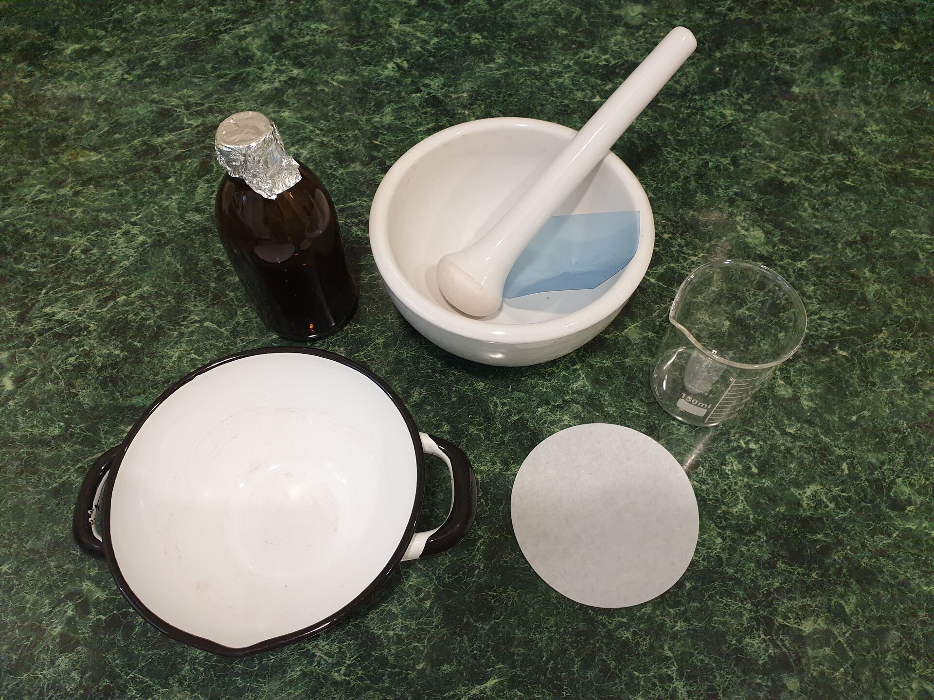 Emulsje. Rycina przedstawia szklaną butelkę z szyjką okrytą folią aluminiową, szklane naczynie z podziałką, mające formę walca, a na górnej krawędzi wyżłobienie, które pomaga w przelewaniu. Ponadto na rycinie są przedstawione: miska, tłuczek (czyli moździerz z pistlem) oraz folia zabezpieczająca, okrągły, papierowy filtr i ceramiczna miska z uchami i wyżłobieniem znajdującym się na górnej krawędzi, ułatwiającym przelewanie. 