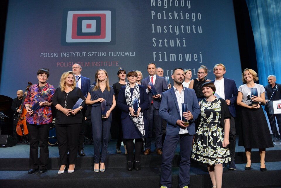 Fotografia przedstawia grupę osób w sile wieku stojących na scenie. Z przodu stoją kobieta i mężczyzna. Kobieta ubrana we wzorzystą, sięgającą za kolana sukienkę. Mężczyzna ma na sobie garnitur i rozpiętą u góry koszulę. W dłoniach trzyma statuetkę w kształcie prostopadłościanu. Postaci stojące na drugim planie trzymają takie same statuetki. W tle logo Polskiego Instytutu Sztuki Filmowej.
