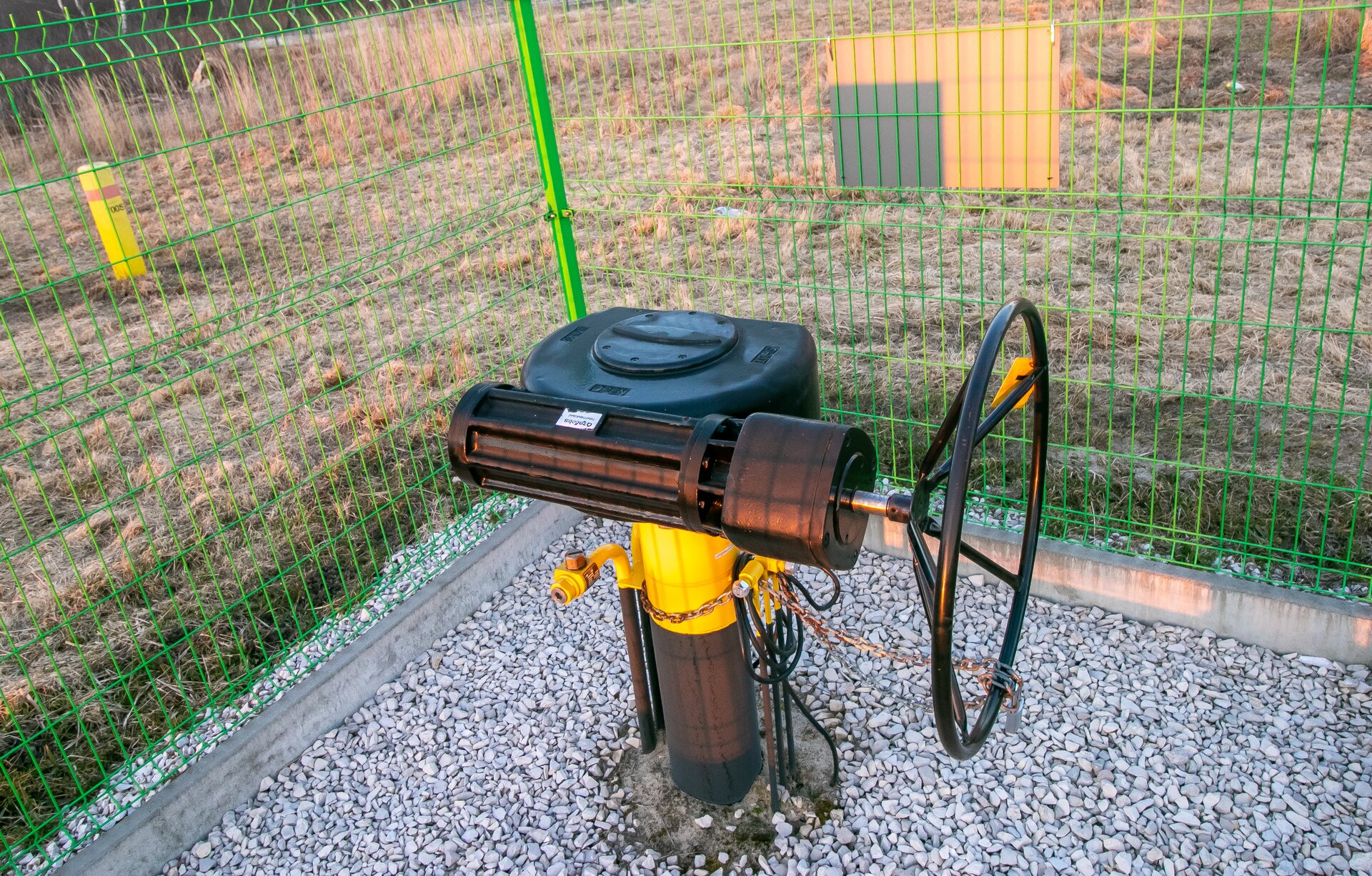 Na zdjęciu widoczna jest zasuwa odcinająca przepływ gazu ziemnego na gazociągu przesyłowym. Zasuwa jest przedstawiona jako rura wychodząca z ziemi. Na końcu rury znajduje się element na którym zamontowany jest kurek o kształcie wielkiego czarnego okręgu. Zasuwa jest ogrodzona.