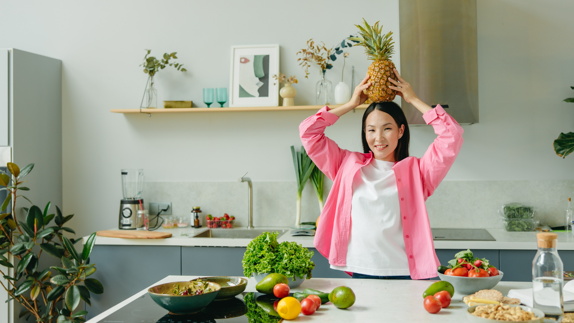 Zdjęcie przedstawia kobietę stojącą przy blacie w kuchni. Kobieta jest uśmiechnięta, na głowie trzyma owoc ananasa. Na blacie leżą owoce i warzywa: sałata, cytryny, pomidory. W tle widoczne są szafki kuchenne, zlew, płyta grzewcza.
