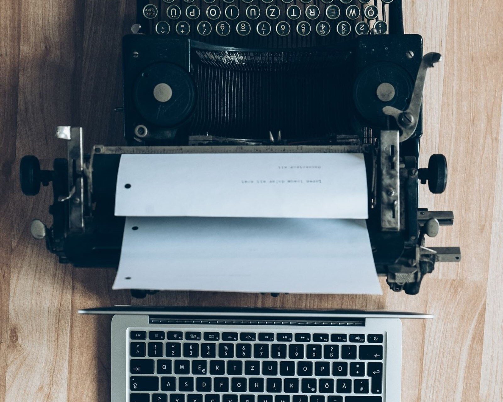 Zdjęcie przedstawia klasyczną maszynę do pisania widzianą z góry, stojącą na drewnianym blacie. W maszynie widoczna jest biała kartka, z zapisanymi dwoma liniami tekstu. Poniżej maszyny do pisania widoczny jest z góry otwarty laptop.