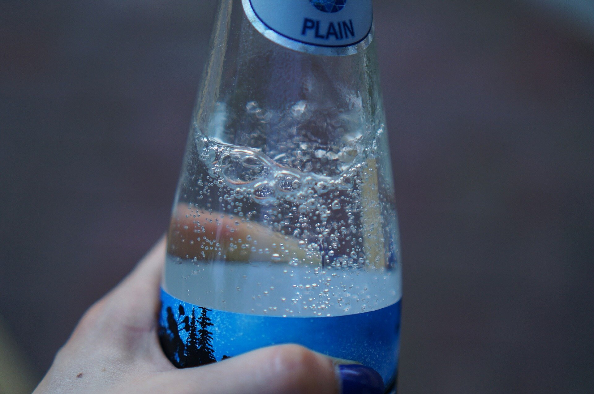 Ilustracja przedstawia fragment szklanej butelki z wodą gazowaną. Widoczny fragment niebieskiej etykiety i mocno musujące pęcherzyki gazu. 