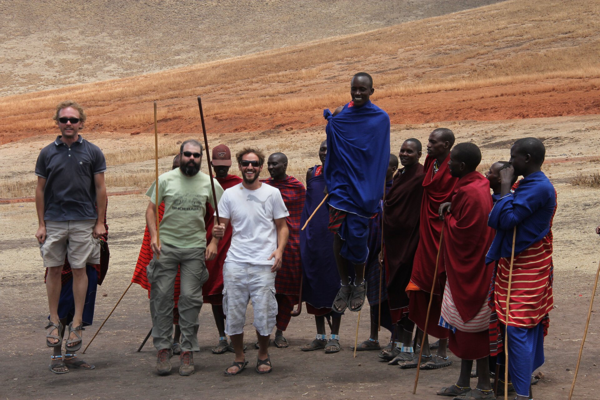 Zdjęcie przedstawia grupę Masajów w tradycyjnych strojach. Jeden z nich wysoko podskakuje. Przed nimi trzech turystów, którzy również uczestniczą w tańcu.