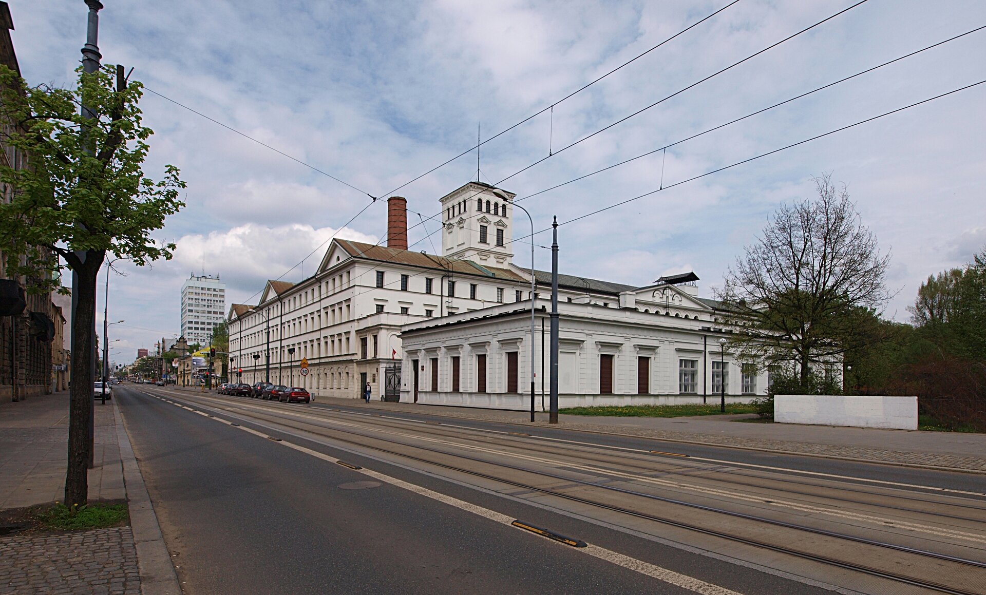 Na zdjęciu szeroka ulica, z namalowanymi pasami, ciągnąca się daleko. Przy niej niewysoki i długi budynek z wysokim kominem. 
