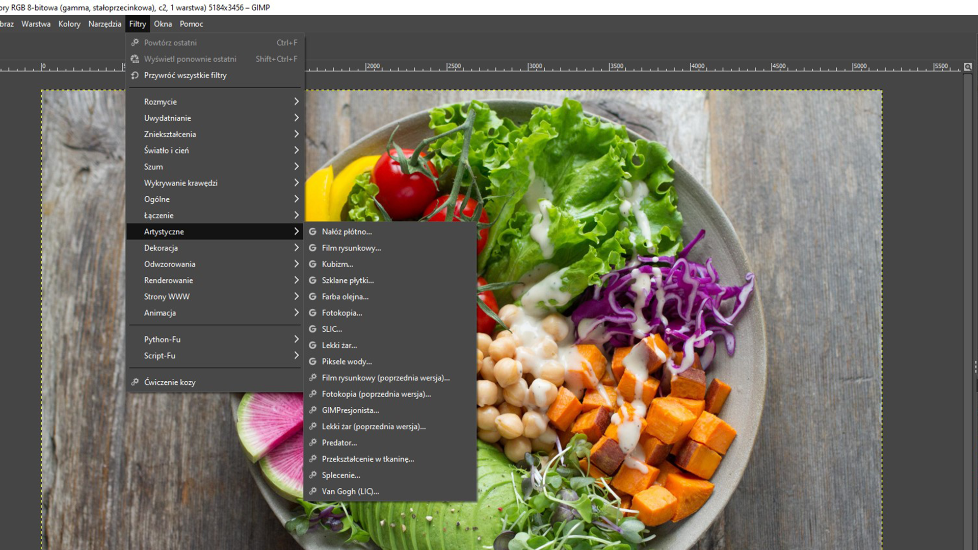 Ilustracja przedstawia okno programu. Z menu głównego wybrano zakładkę Filtry. Z listy wybrano opcję: Artystyczne. Przy Artystyczne lista z różnymi opcjami, na przykład: Nałóż płótno, Film rysunkowy, Kubizm, Szklane płytki, Piksele wody, Splecenie. W tle zdjęcie warzyw leżących na talerzu. 