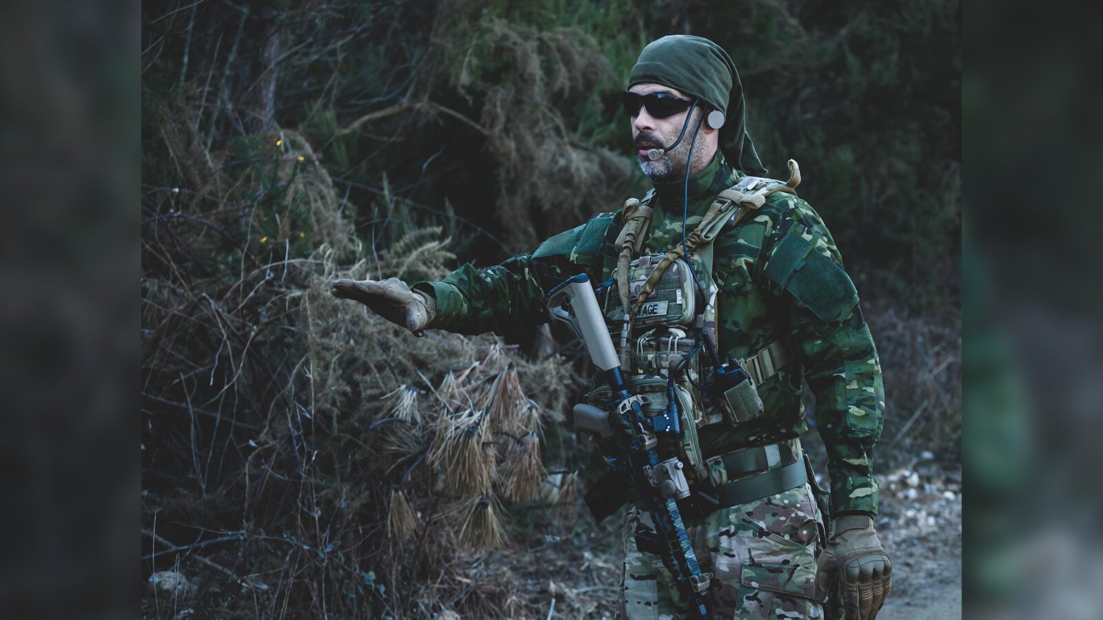 Zdjęcie przedstawia osobę ubraną w maskujący strój i mającą słuchawki z mikrofonem przy ustach. 