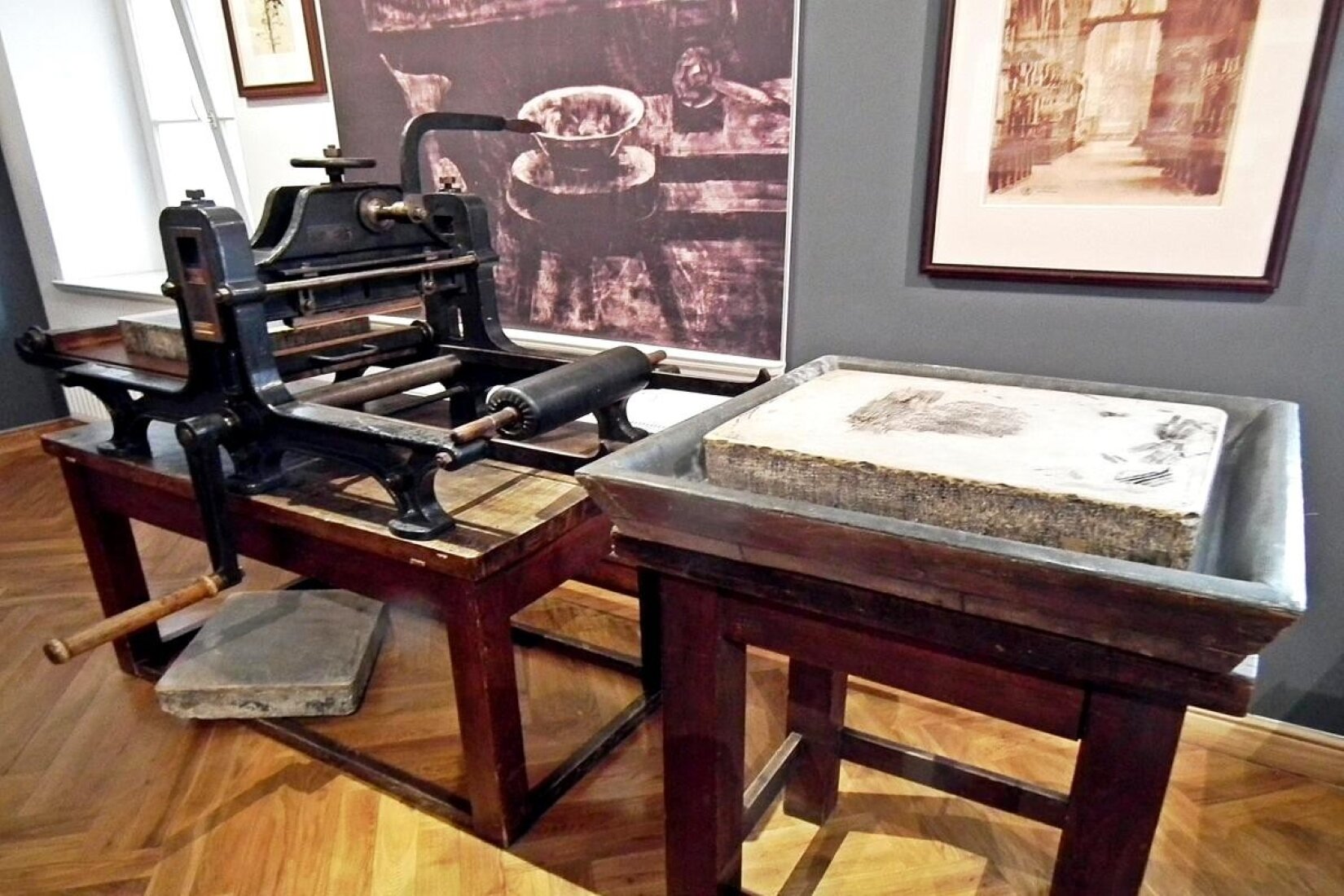 Fotografia przedstawia rekonstrukcję pracowni graficznej Leona Wyczółkowskiego, w której znajduje się prasa i wanna litograficzna. Prasa kształtem przypomina czworonożny drewniany stół, na którym umieszczone zostały metalowe rolki oraz obciążniki. Jej podstawa wykonana jest z bordowego drewna, a samo narzędzie jest czarne, z drewnianymi elementami. Poniżej blatu narzędzia jest długa jasnobrązowa rączka. Przy podstawie umieszczono jasny, prostokątny kamień, leżący częściowo na drewnianej podłodze o ciepłym odcieniu ze wzorem w jodełkę, a częściowo na deskach prasy. Wanna litograficzna, znajdująca się na prawo od prasy, również została ułożona na bordowej drewnianej podstawie. Jest prostokątna oraz szara. Jej brzegi odchylają się nieznacznie do zewnątrz. Umieszczono w niej duży, prostokątny kamień o nieregularnej strukturze oraz szarym odcieniu. Oba narzędzia stoją na podłodze. Z tyłu, na ścianie, wiszą dwa obrazy – jeden przedstawia wnętrze zacienionego pomieszczenia, w którym stoi stołek oraz umieszczone na nim naczynie, a drugi jasną przestrzeń ograniczoną ścianami. Z lewej strony jest uchylone okno, wpuszczające do pomieszczenia światło, oraz fragment ciemnej ściany.