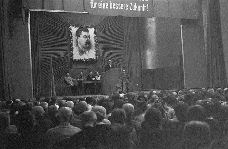 Zdjęcie przedstawia scenę, przed którą siedzi tłum ludzi. Są skupieni na tym, co się dzieje na scenie. Na niej przy mównicy stoi przemawiający mężczyzna. Pod mównicą stoi symbol sierpa i młota. Z tyłu przy stole siedzi grupa mężczyzn. Nad nimi wisi duży portret Józefa Stalina otoczony kwiatami. Wyżej widoczny jest napis w języku niemieckim.