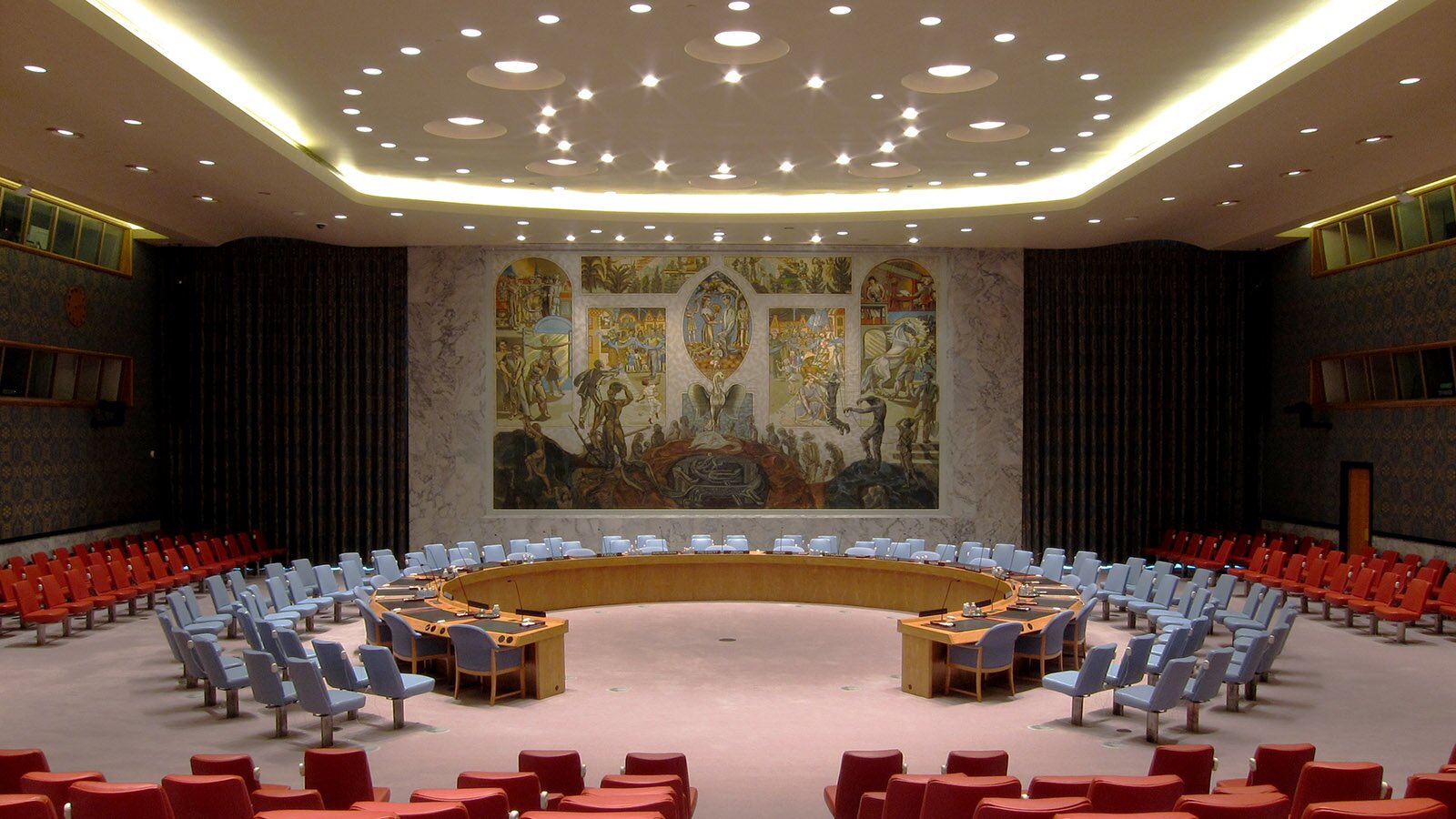 Zdjęcie przedstawia salę obrad, w której pośrodku widoczny jest stół w kolorze jasnego drewna, ustawiony w formie otwartego okręgu. Po zewnętrznej stronie stołu widoczne są niebieskie fotele. Przy każdym miejscu przy stole widoczny jest mikrofon. Wokół stołu widoczne są dwa rzędy krzeseł obitych niebieskim materiałem. W niewielkiej odległości od nich widoczne są kolejne rzędy foteli w czerwonym obiciu, które dochodzą do ścian pomieszczenia. Pośrodku na ścianie widoczne jest renesansowe malowidło. Pod podwieszanym sufitem widoczna jest instalacja z wieloma lampkami. Na podłodze widoczny jest dywan w kolorze brudnoróżowym.