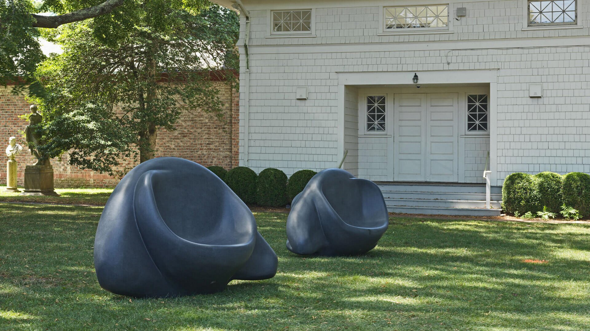 Zdjęcie przedstawia instalację artystyczną umieszczoną na trawie w parku. „Oczy - ławki” to dwa elementy wykonane z granitu, w ciemnoszarym kolorze. Na tym ujęciu ławki zostały przedstawione z tyłu. Wyżłobione zostało siedzisko. Powierzchnia materiału jest gładka. Ławki znajdują się w pewnej odległości od siebie. W tle stoi biały budynek z dużymi drzwiami i kwadratowymi okienkami, a także drzewa, krzewy i dwie inne rzeźby. Fotografia została wykonana w słoneczny dzień.