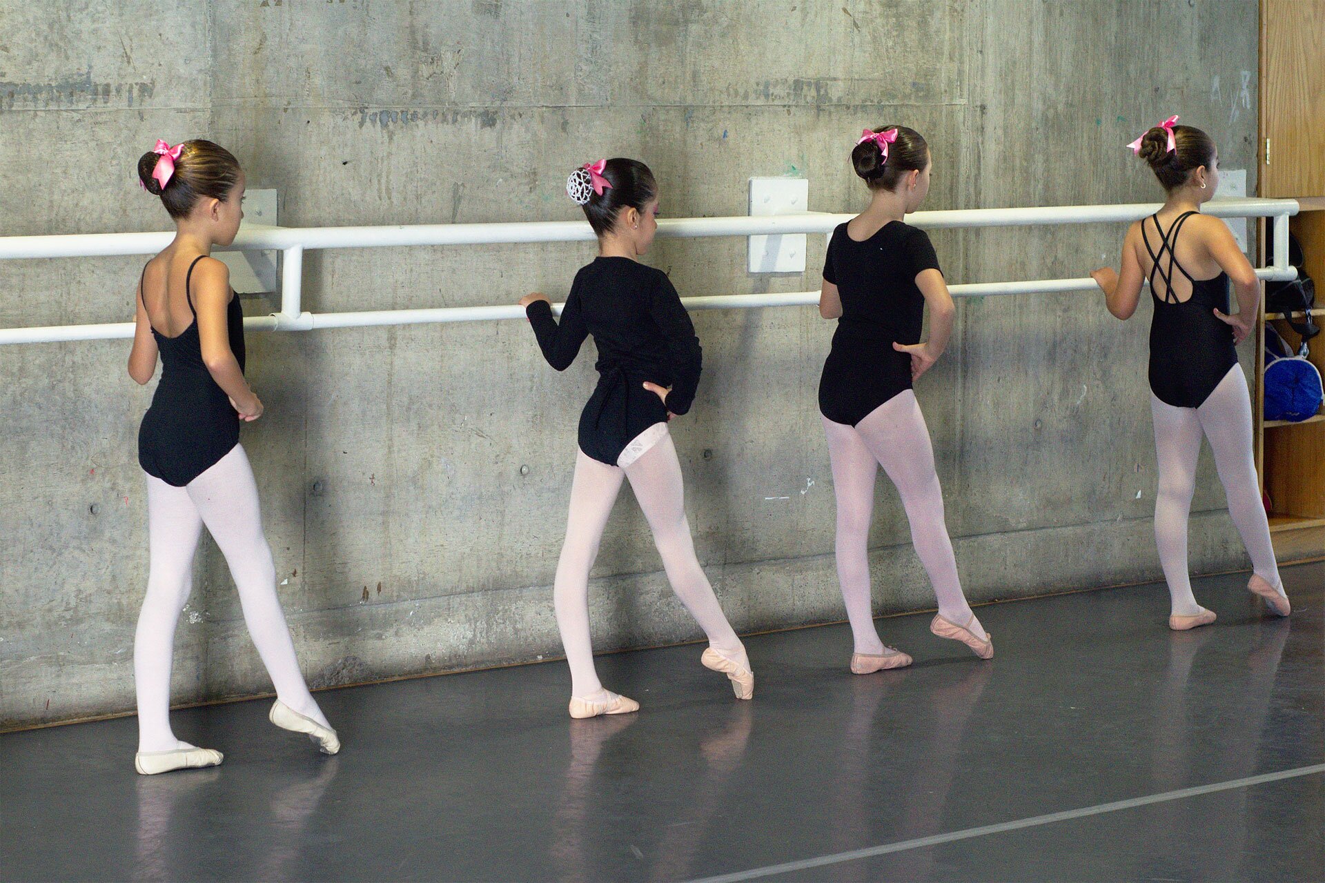 Fotografia przedstawia cztery dziewczynki, podczas zajęć baletowych, stojące bokiem przy ścianie przy białej, podwójnej poręczy. Prawe dłonie oparte na biodrach, prawe nogi wysunięte do przodu, palce stóp obciągnięte. Patrzą wyprostowane przed siebie. Dziewczynki są ubrane w czarne body i białe rajstopy. Na stopach jasne baletki. Na głowie okrągłe koki ozdobione różowymi kokardami.