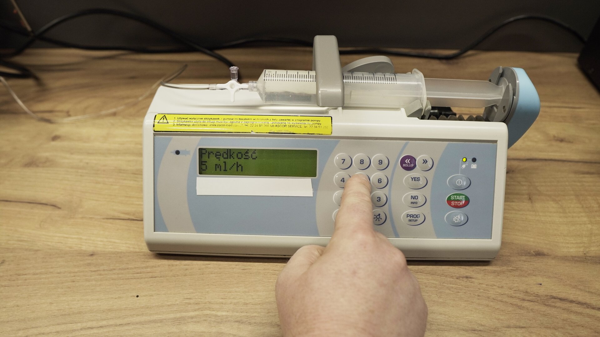 Na zdjęciu strzykawka wypełniona wodą znajduje się w ramieniu pompy infuzyjnej. Na ekranie aparatu wyświetlana jest informacja: "Prędkość 5 ml/h". Technik wciska przycisk 5 na obudowie aparatu. 