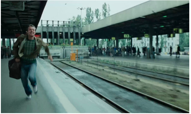 Fotografia przedstawia mężczyznę w sile wieku. Biegnie wzdłuż peronu kolejowego. Postać ukazuje owalną twarz. Mężczyzna ma wysokie czoło, zaciśnięte powieki, duży nos i grymas na ustach. Odsłania zęby. Jest ubrany w kurtkę, sweter w kratę oraz dżinsowe spodnie. Na prawym ramieniu ma zawieszoną dużą, skórzaną torbę. Na innym peronie ukazanym po prawej stronie stoją pasażerowie. Kierują się w przeciwną stronę, prawdopodobnie ku tunelowi. W tle jasne niebo.