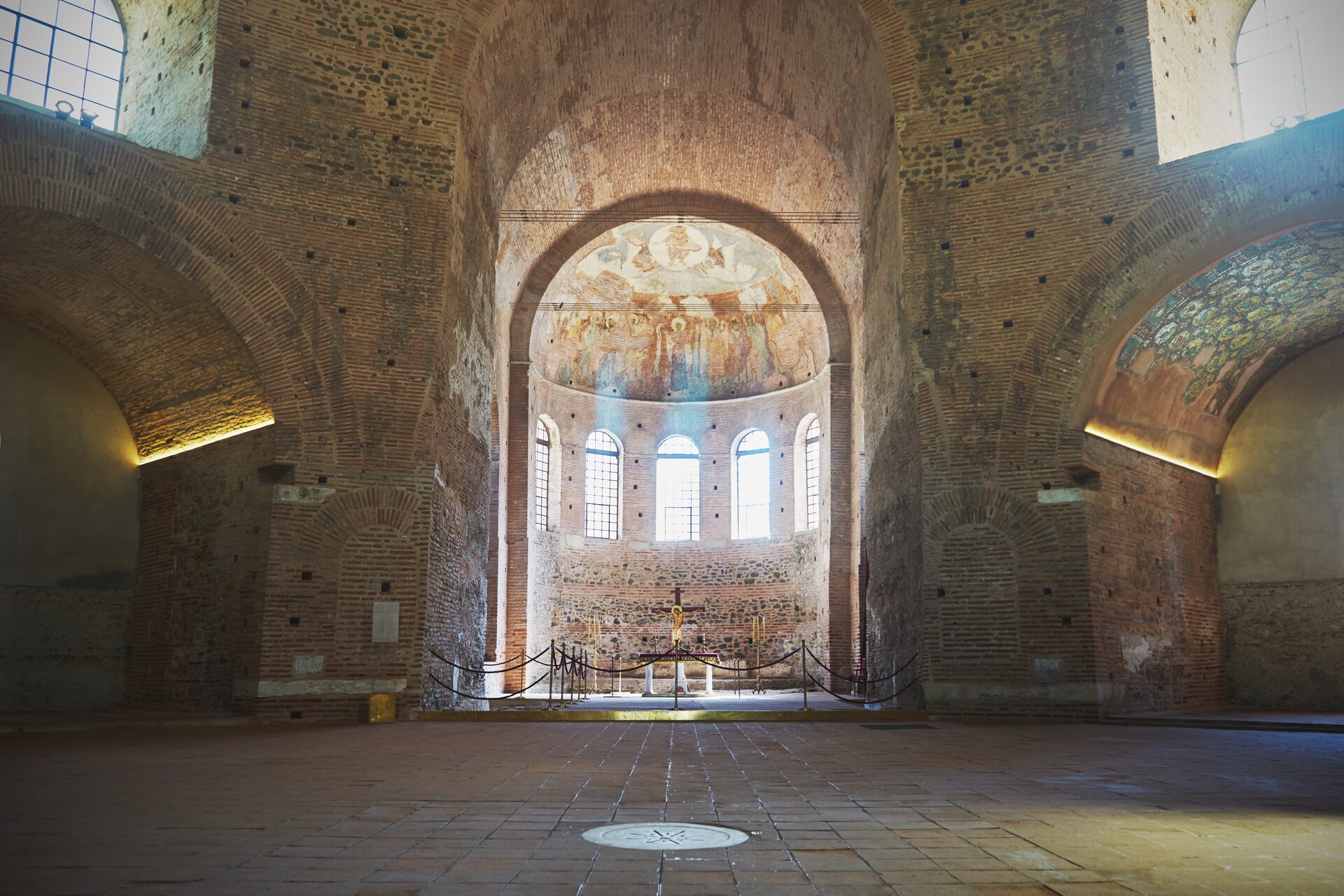 Ilustracja przedstawia wnętrze rotundy z IV wieku znanej jako kościół Agios Georgios. Budynek wykonano głównie z cegły i kamienia. W centralnej części znajduje się krzyż, zabezpieczony balustradą przed zwiedzającymi. Krzyż jest umieszczony we wnęce, części rotundy zwanej apsydą, z pięcioma wysokimi oknami, zwieńczonej półkolistym sklepieniem. Po lewej i prawej stronie znajdują się niższe sklepienia zdobione mozaikami.