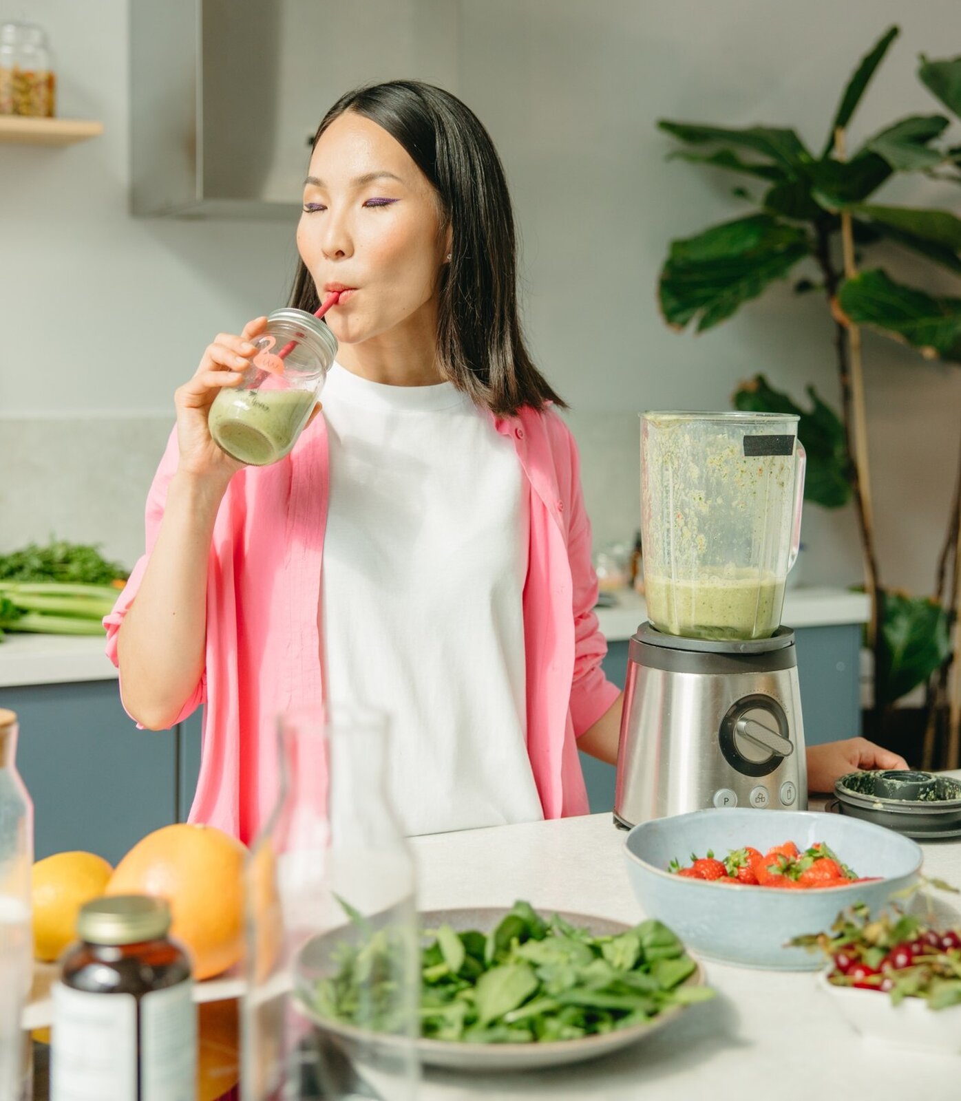 Zdjęcie przedstawia młodą Azjatkę pijącą ze smakiem warzywno‑owocowy koktajl. Kobieta znajduje się w kuchni, przy stole, na którym znajduje się blender kielichowy, wypełniony częściowo zielonkawym koktajlem. Ponadto na stole stoi miska z truskawkami, miska z czereśniami, miska ze szpinakiem. Nieopodal nich leżą pomarańcza i grejpfrut.