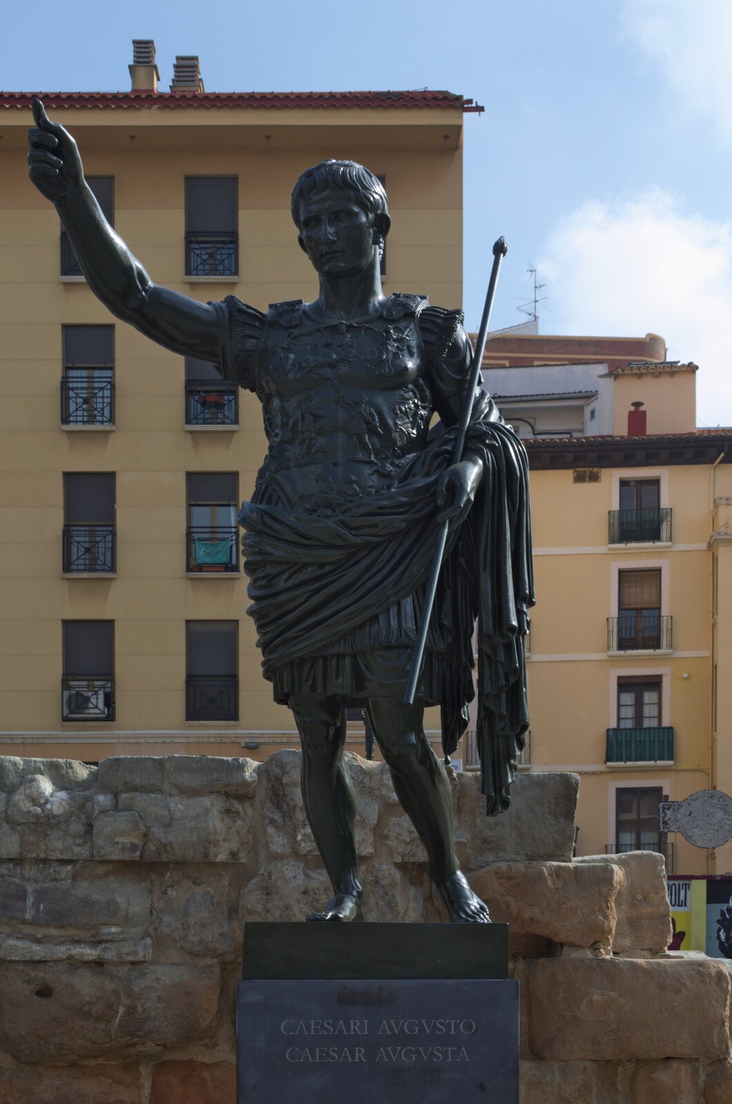 Fotografia przedstawia pomnik z ciemnego metalu uwieczniający mężczyznę, który patrzy przed siebie. Trzyma w lewej dłoni przedmiot przypominający włócznię, a prawą dłoń kieruje ku górze, z uniesionym palcem wskazującym. Lewą nogę ma wysuniętą do tyłu; opiera ją na palcach dla równowagi. Ma na sobie krótką szatę, której część jest przerzucona na lewą rękę, jest bosy oraz posiada bardzo bogato zdobioną zbroje. Tło stanowią jasnożółte budynki oraz fragment kamiennego muru. Na podwyższeniu, na którym umieszczono pomnik, obecne są napisy „CAESARI AVGVSTO” oraz „CAESAR AVGVSTA”.