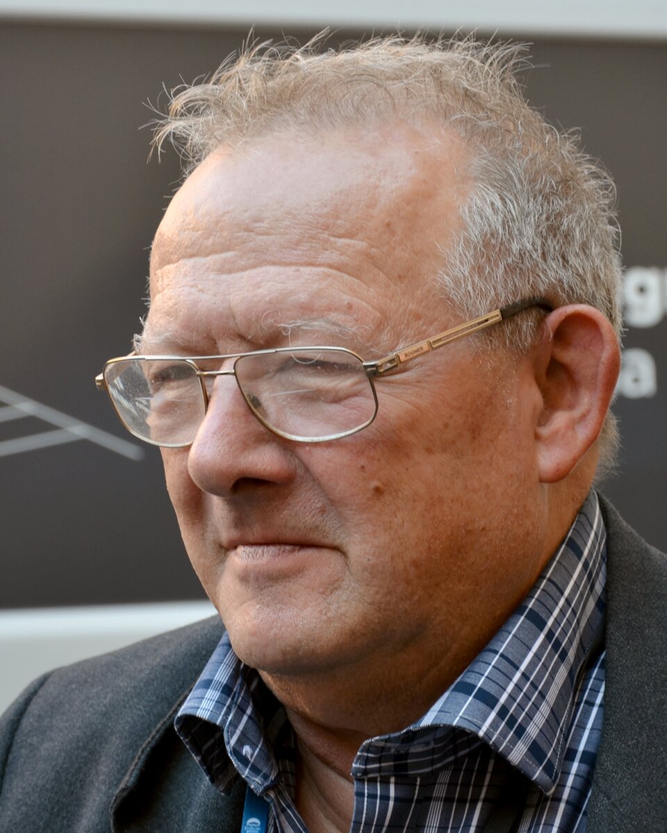 Zdjęcie przedstawia starszego mężczyznę. Jest zwrócony twarzą w lewą stronę zdjęcia. Ma krótkie, przerzedzone, siwe włosy, zmarszczki na twarzy, małe usta oraz okulary w cienkich oprawkach. Ubrany jest w szarą marynarkę oraz niebieską koszulę w kratę.