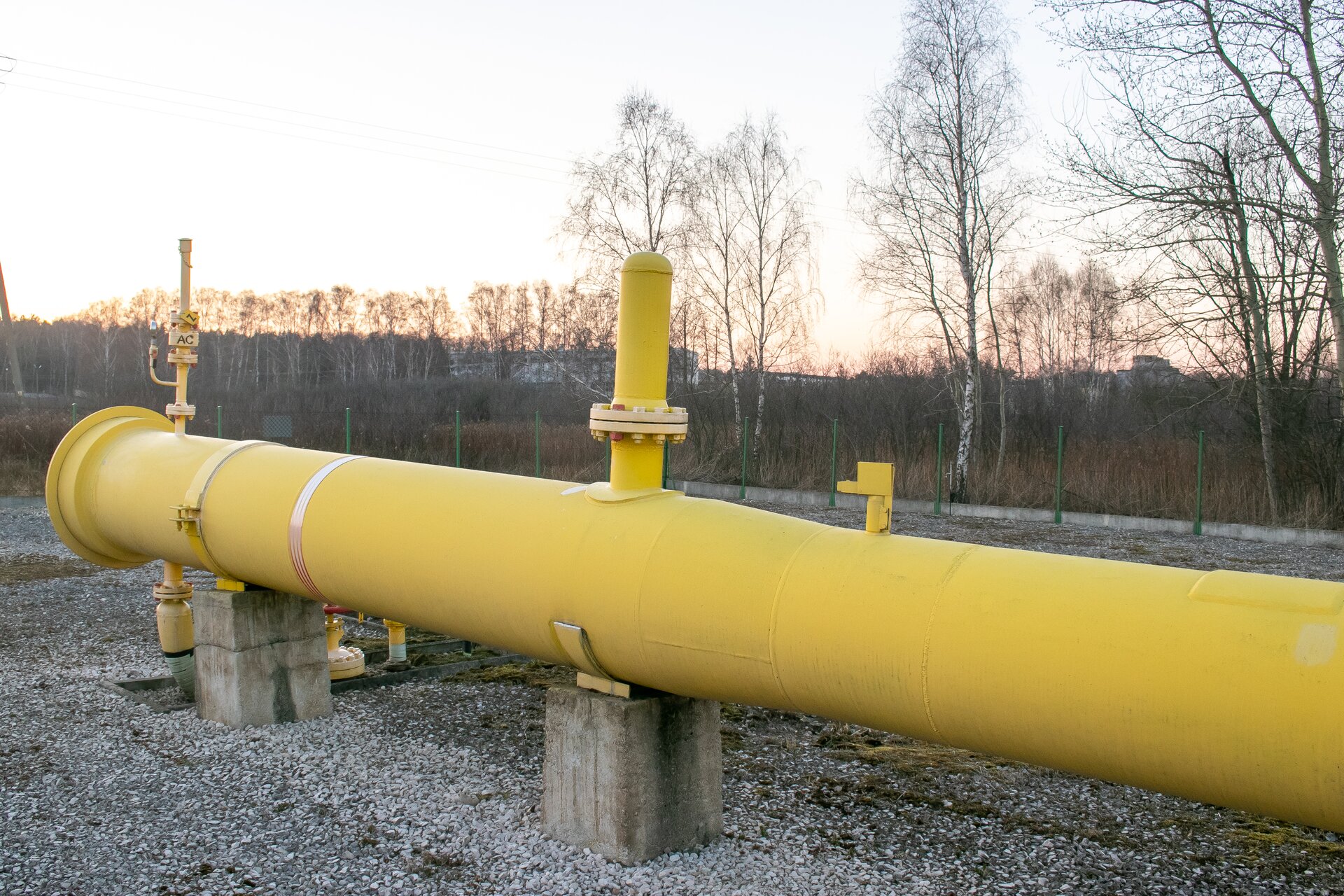 Na zdjęciu przedstawiony jest fragment rurociągu gazowego. Przekrój rurociągu ma kształt okręgu. Rurociąg wykonany jest ze stali pomalowanej na żółty kolor. Na górze rurociągu widać dwa elementy rurowe które są prostopadłe do niego. Rurociąg posadowiony jest na betonowych prostopadłościennych klockach. Klocki znajdują się na podłożu na którym wysypany jest żwir.