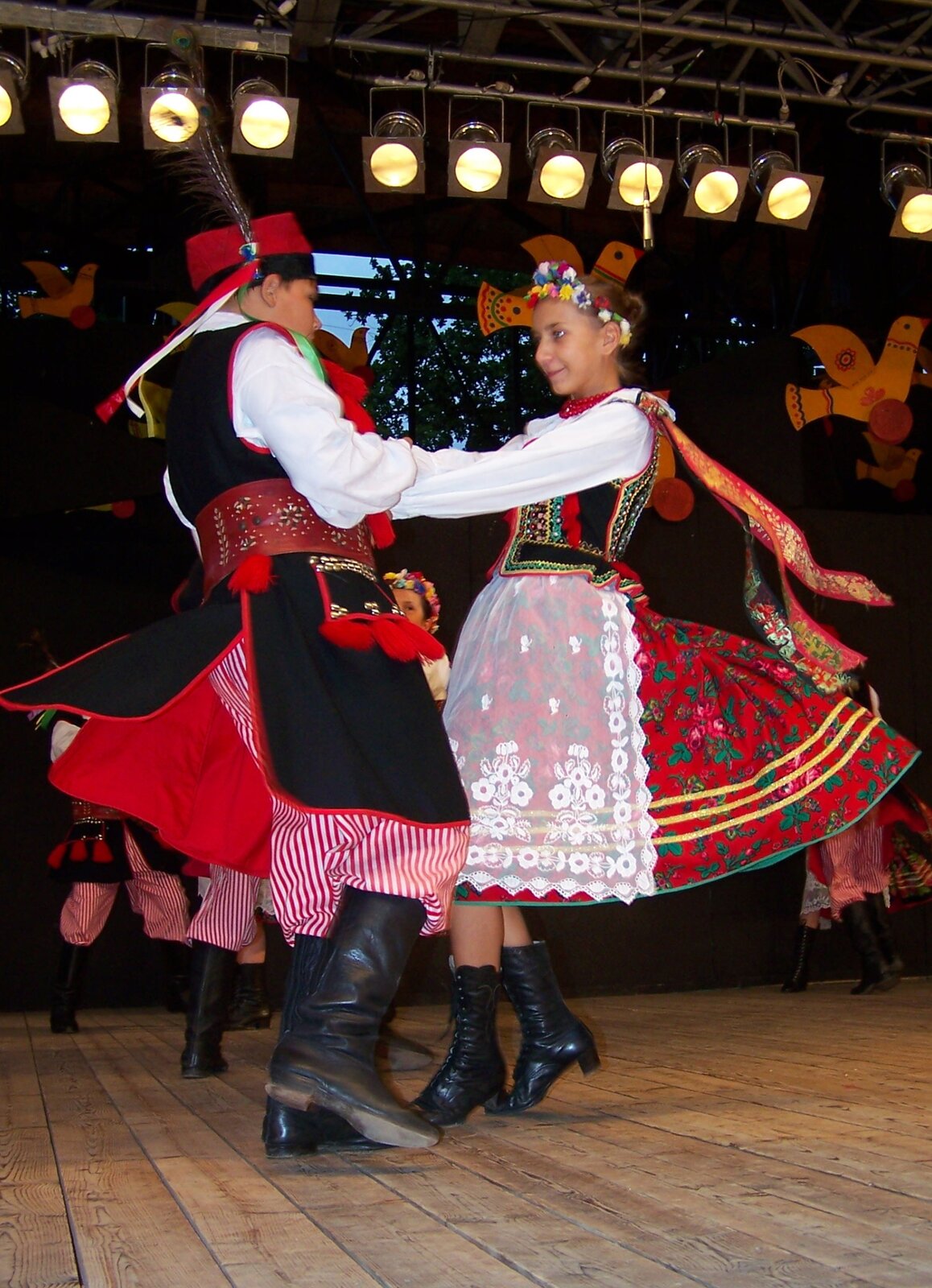 Ilustracja przedstawia prezentacje tradycyjnego stroju krakowskiego. Na ilustracji znajduje się tańcząca para. Chłopiec ubrany jest w kozaki, szerokie biało‑czerwone spodnie w paski, długą czarną kamizelkę oraz białą koszulę. Na głowie chłopca znajduje się kapelusz z przyczepionym piórem. Dziewczynka ubrana jest w czarne kozaki, czerwoną suknie z kwiatowym wzorem, białą koszulę. Na głowie dziewczynki znajduje się wianek z kwiatów. W tle fotografii znajdują się inne tańczące pary. 