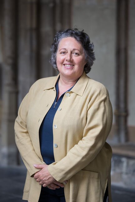 Zdjęcie przedstawia dojrzałą kobietę. Ma owalną twarz, ciemnosiwe, lekko kręcone, krótkie włosy. Jest uśmiechnięta. Trzyma się ręce przed sobą. Ubrana jest w niebieską koszulę i jasny sweter.