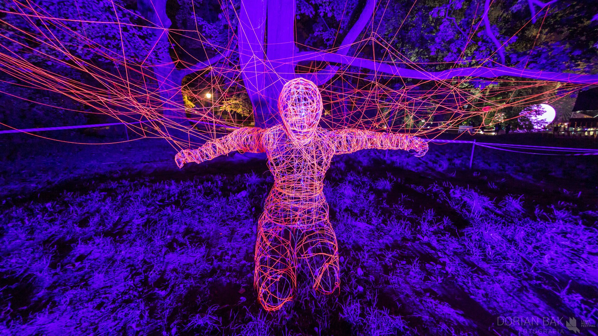 Na zdjęciu znajduje się instalacja z drutu przedstawiająca klęczącą postać ludzką z rozłożonymi ramionami. Za postacią jest pajęczyna z drutu. Postać, jak i pajęczyna podświetlone są kolorem pomarańczowym. Instalacja znajduje się na trawniku, na tle drzew, które są podświetlone fioletowym światłem. Tło stanowi czarne nocne niebo, fotografia jest ciemna. 