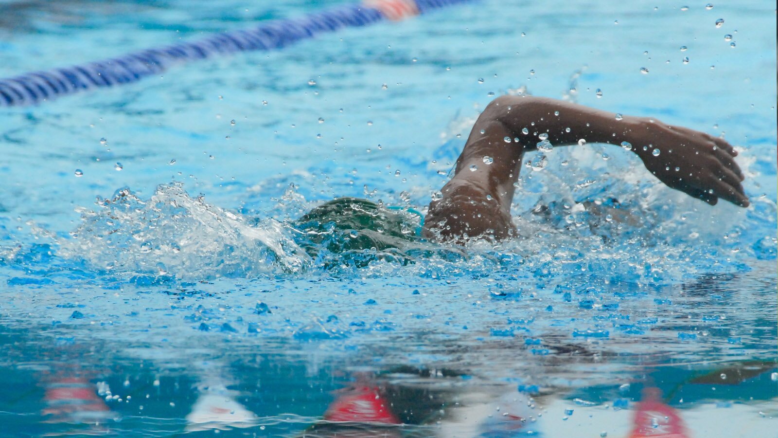Zdjęcie przedstawia pływaka płynącego w basenie. Widoczna jest lewa ręka wystająca ponad taflę wody oraz zanurzona pod wodą głowa. W tle widoczna jest linia złożona z ciemnoniebieskich elementów, która wyznacza tor pływacki.