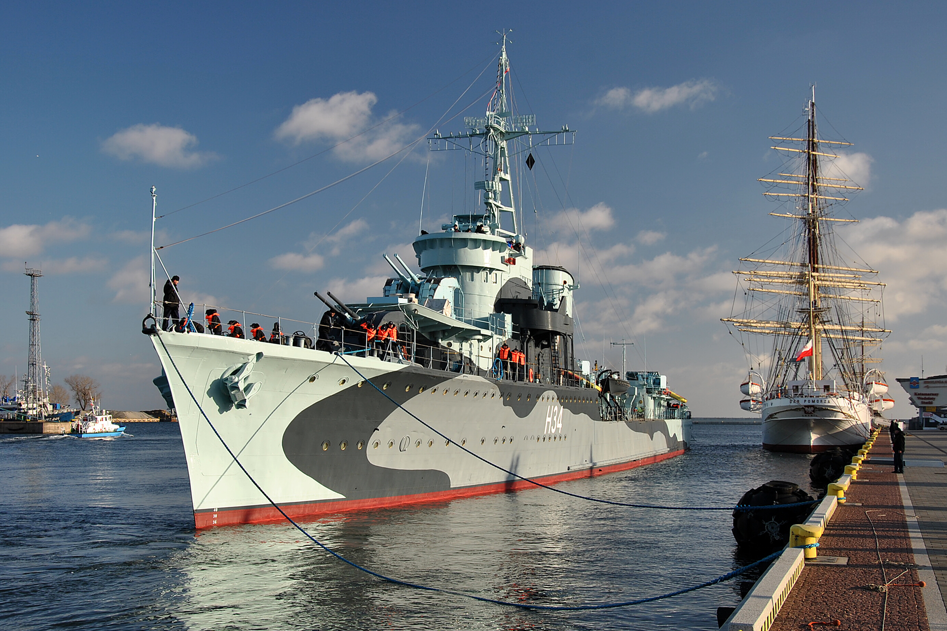 Fotografia przedstawiająca okręt-muzeum "Błyskawica" dobijający do nabrzeża w Gdyni. Za nim zacumowany trzymasztowiec statek-muzeum "Dar Pomorza". Niebo niebieskie z nielicznymi białymi chmurami. 