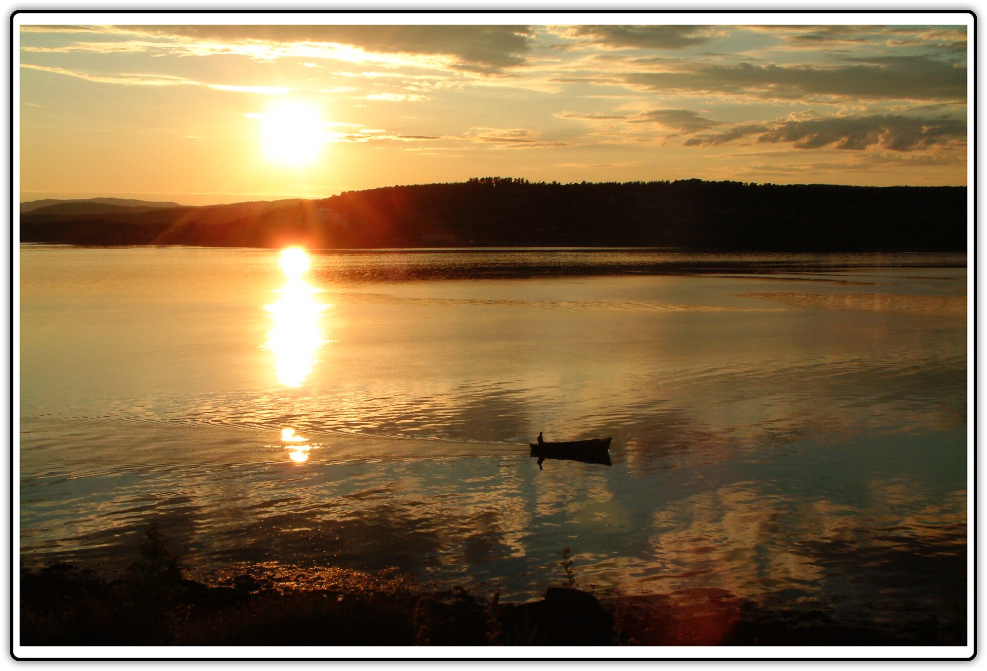 Fotografia przedstawiająca drewnianą łódkę płynącą po jeziorze o zmierzchu na tle zachodzącego słońca. W łódce  tyłem siedzi długowłosa osoba. W oddali widać zarys zacienionego lądu.