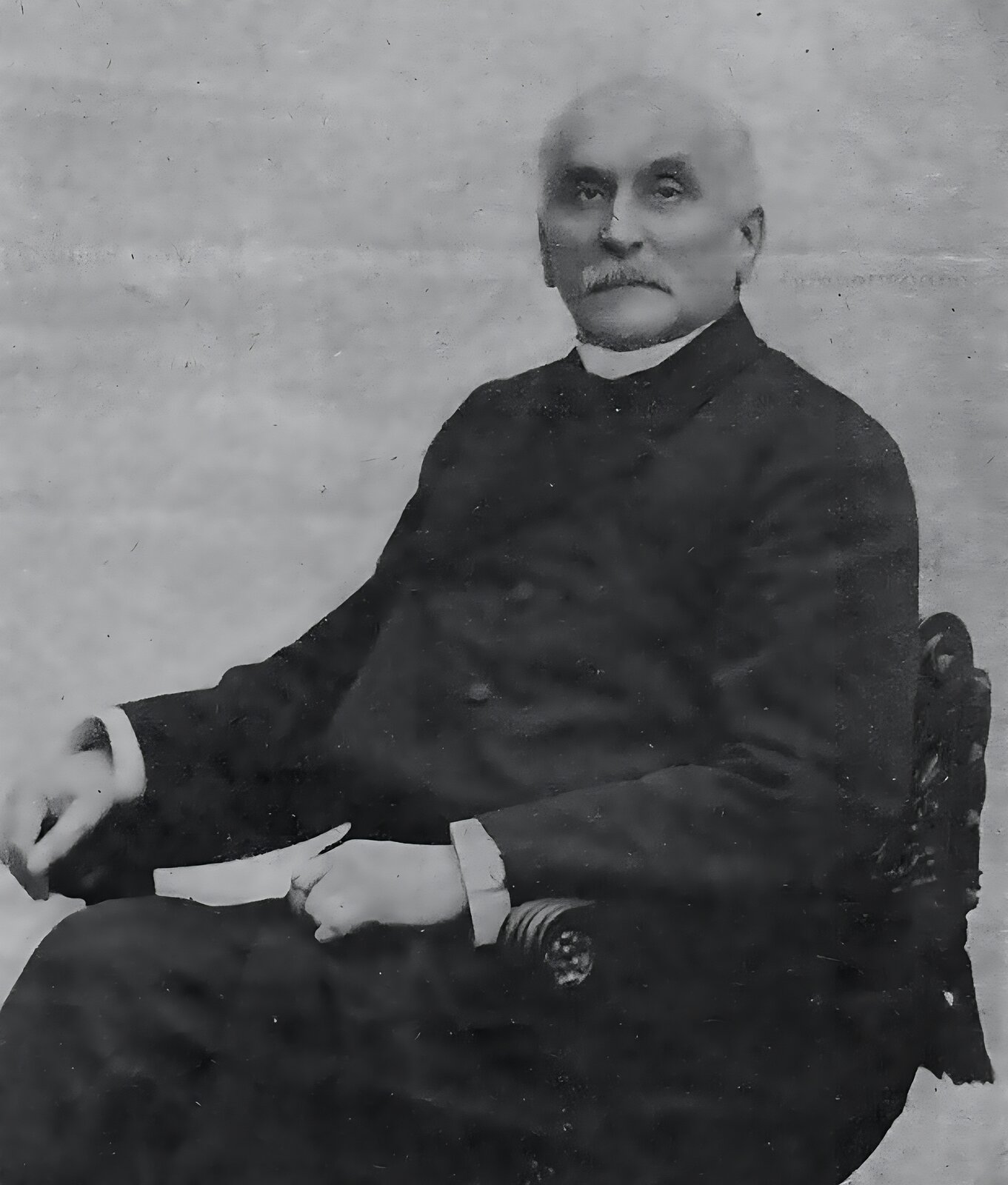 Zdjęcie archiwalne przedstawia starszego mężczyznę siedzącego na krześle. Nosi on czarny garnitur, szyję oplata biały kołnierz bez guzika. Z rękawów marynarki wystają mankiety białej koszuli. Mężczyzna ma gęste, siwe wąsy, włosy widoczne jedynie powyżej obu uszu. Oczy ma otwarte i zwrócone w kierunku widza. Krzesło stylizowane zgodnie z modą przełomu XIX i XX wieku, z finezyjnie wykończonym oparciem i podłokietnikiem. 