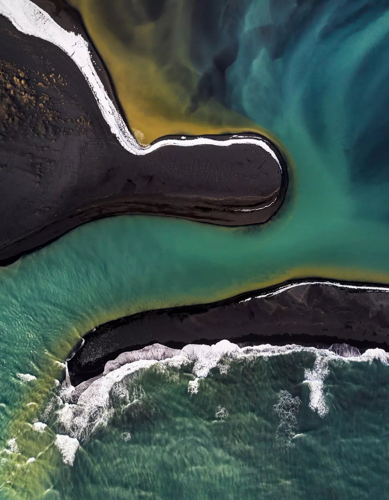 Fotografia, wykonana z lotu ptaka, przedstawia widok na dwie, czarne zatoki lub wyspy oraz powierzchnię wody. Zatoka uchwycona wyżej jest szersza, a na jej końcu można zauważyć że jest w kształcie przypominającym owal. Przy jej górnej krawędzi biegnie biała linia, a po lewej stronie widoczne są jaśniejsze miejsca, przypominające brązowe skały. Zatoka przedstawiona niżej, poniżej centrum fotografii, jest węższa, bardziej podłużna. Przy jej górnej krawędzi również widoczny jest biały pas, jednak rozpoczyna się on bliżej jej środka i ciągnie się ku prawemu końcowi. Tuż przy zatokach woda ma odcień brązowo‑żółty, następnie przechodząc w turkus oraz kolor niebieski. Poniżej mniejszej zatoki widoczna jest też biała powierzchnia przypominająca wzburzoną wodę.