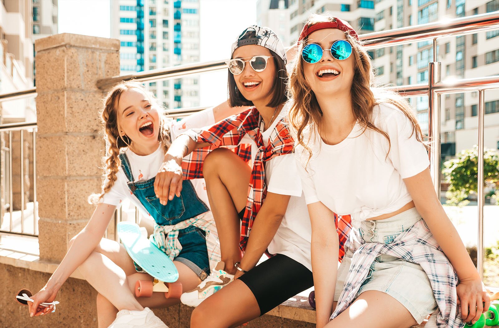 Zdjęcie przedstawia trzy uśmiechające się nastolatki siedzące na murku. Dziewczyna od lewej strony ma długie, jasne włosy zaplecione w warkocze. Jest ubrana w białą bluzkę i dżinsowe ogrodniczki. Jest zwrócona w prawą stronę, lewą ręką dotyka głowy dziewczyny siedzącej po jej lewej stronie, zaś prawą rękę, w której trzyma okulary przeciwsłoneczne, ma opartą na podciągniętej nodze. Dziewczyna w środku ma półdługie, ciemne włosy, na które założyła odwróconą czapkę z daszkiem, a na twarzy ma okulary przeciwsłoneczne. Jest ubrana w białą bluzkę z krótkim rękawem, krótkie, czarne leginsy, a na szyi ma przewiązaną koszulę w kratę. Prawą nogę ma podciągniętą i opartą na murku, a na niej położoną prawą rękę. Dziewczyna po prawej stronie ma długie, jasne włosy, na które założyła odwróconą czapkę z daszkiem, a na twarzy ma okulary przeciwsłoneczne. Jest ubrana w białą bluzkę z krótkim rękawem, dżinsową spódnicę, a w pasie ma przewiązaną koszulę w kratę. Rękami opiera się na murku. W tle, za barierką, widoczne są wysokie budynki. 