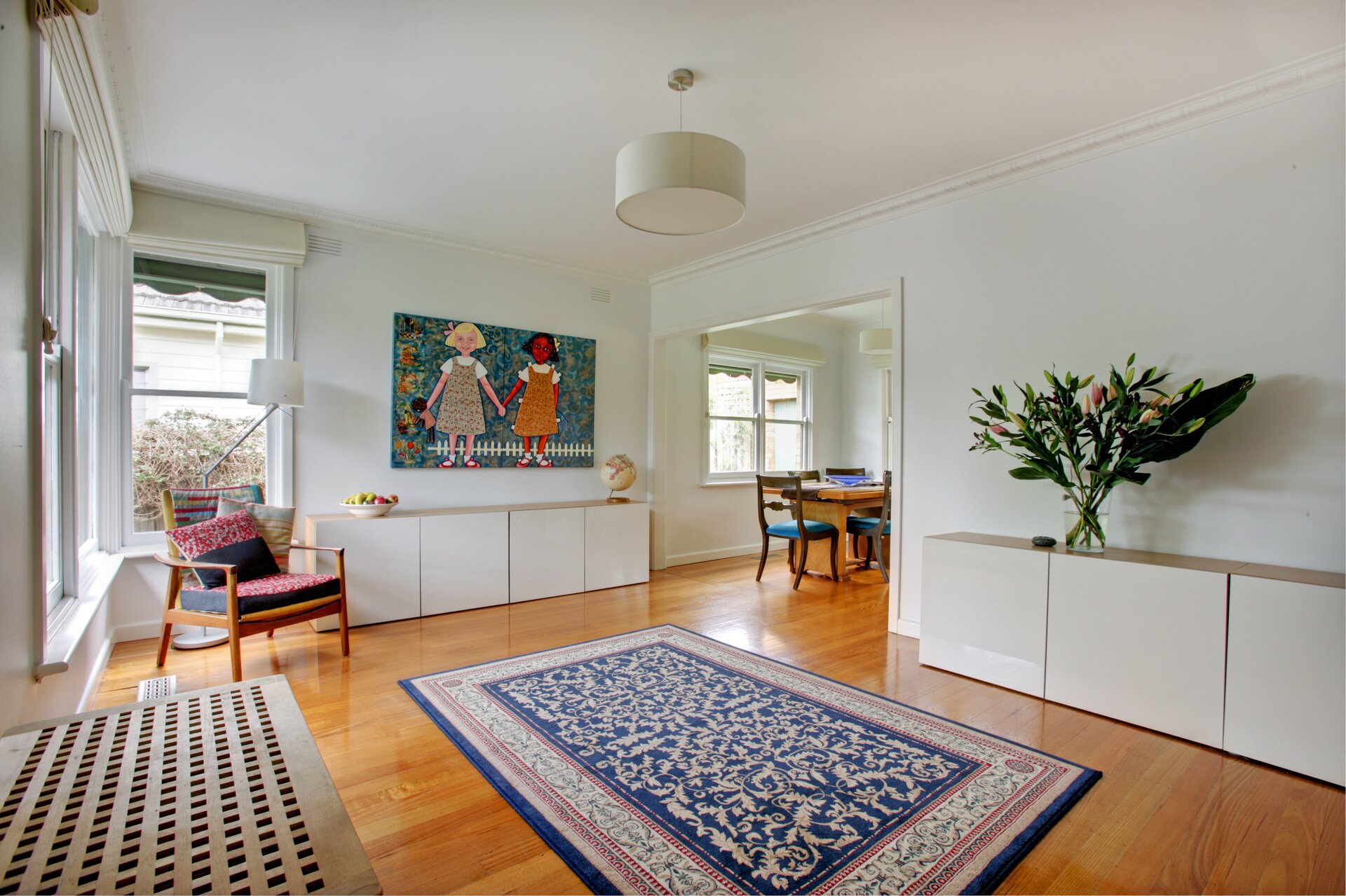 Fotografia przedstawia pokój. W pomieszczeniu dominuje kolor biały. Kolorowe elementy to: obraz dwóch trzymających się za ręce dziewczynek wiszący na wprost, stojący pod oknem drewniany fotel, niebieski dywan we wzory (ornamenty kwiatowe) leżący na podłodze oraz zielony bukiet kwiatów stojący w wazonie na komodzie. W rogu pokoju znajduje się wejście do kolejnego pomieszczenia, w którym widoczne są krzesła z niebieskimi obiciami i stół. 