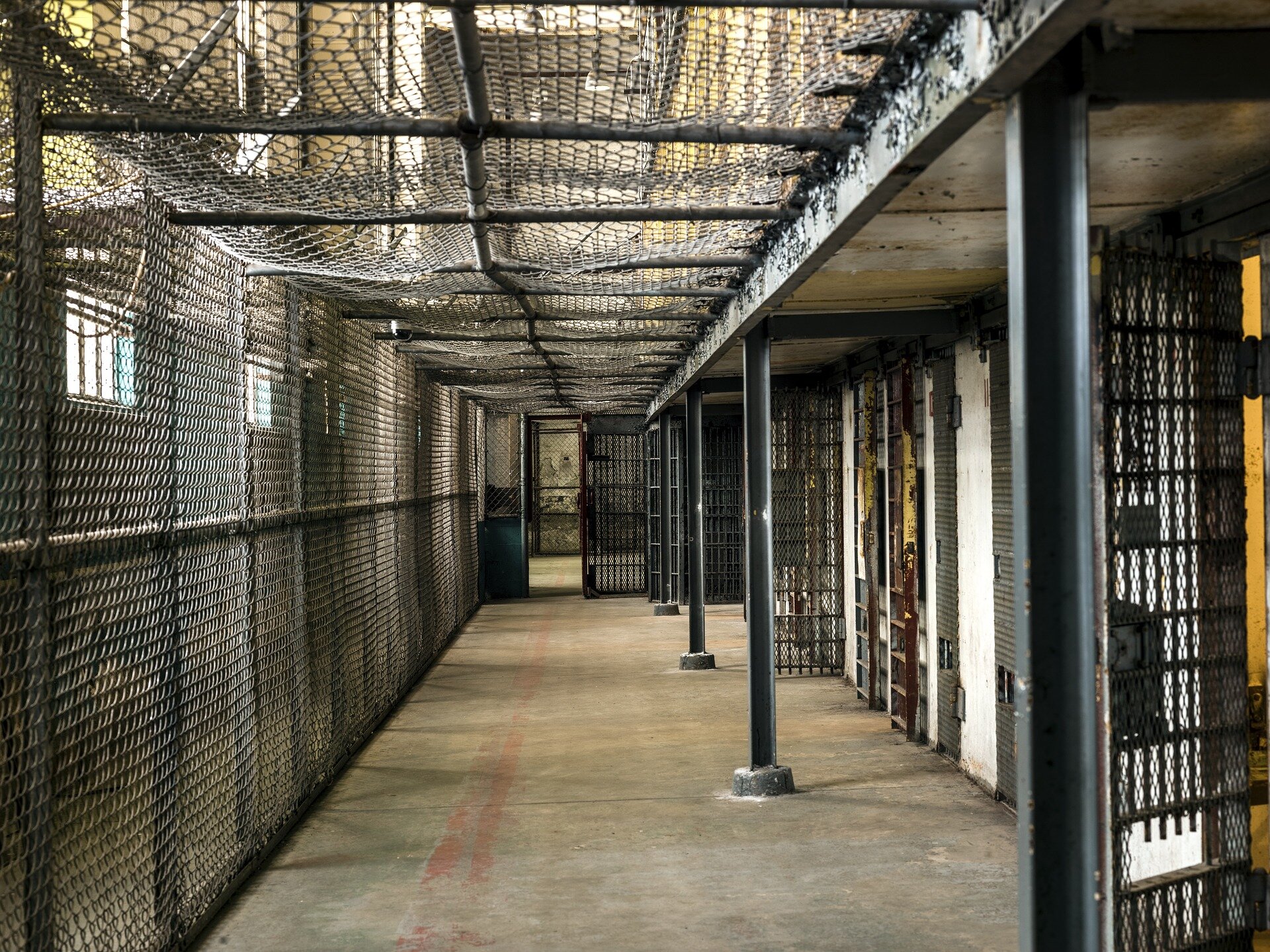 Zdjęcie przedstawia wnętrze więziennego korytarza. Po prawej stronie znajdują się wejścia do cel. Korytarz zabezpieczony jest z lewej strony i od góry gęstą siatką. 