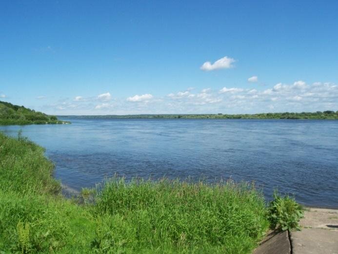  Na zdjęciu rzeka. Jej koryto jest szerokie a woda spokojna. Na obu brzegach rośnie trawa